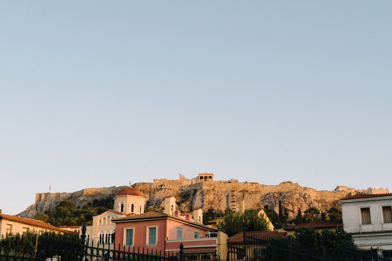 Ancient Acropolis visible atop a hill with colorful buildings in the foreground at sunset.