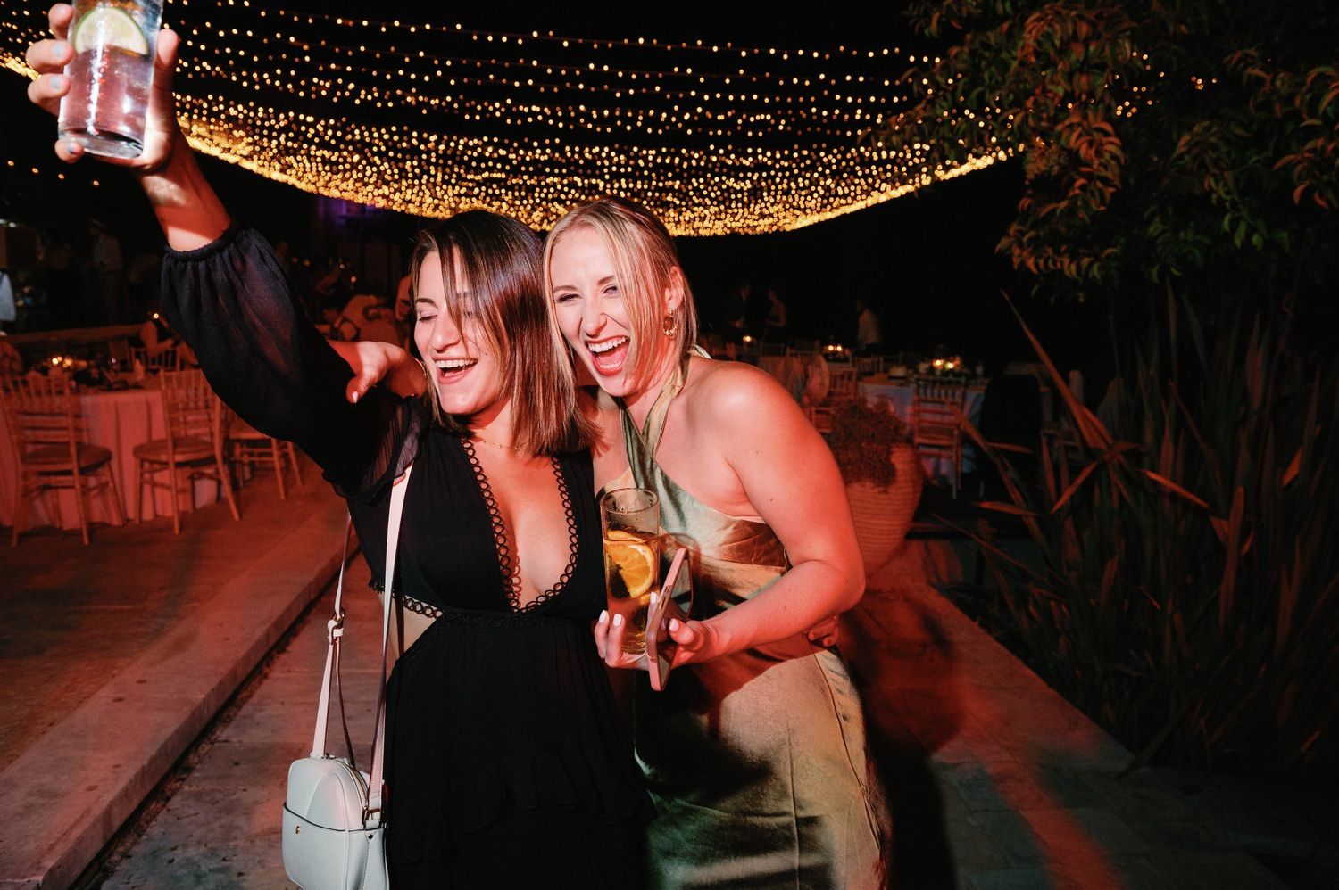 Two party guests taking a selfie together under string lights at an outdoor celebration.