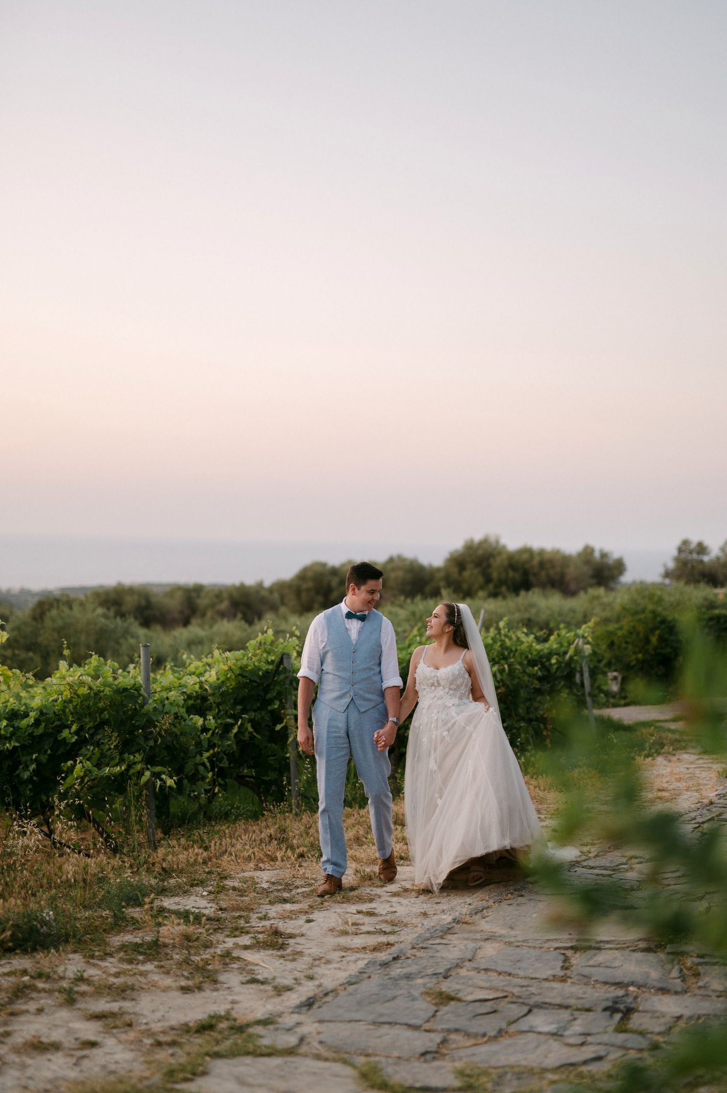 A couple walks hand in hand through a vineyard at sunset on a stone path wearing formal wedding attire.