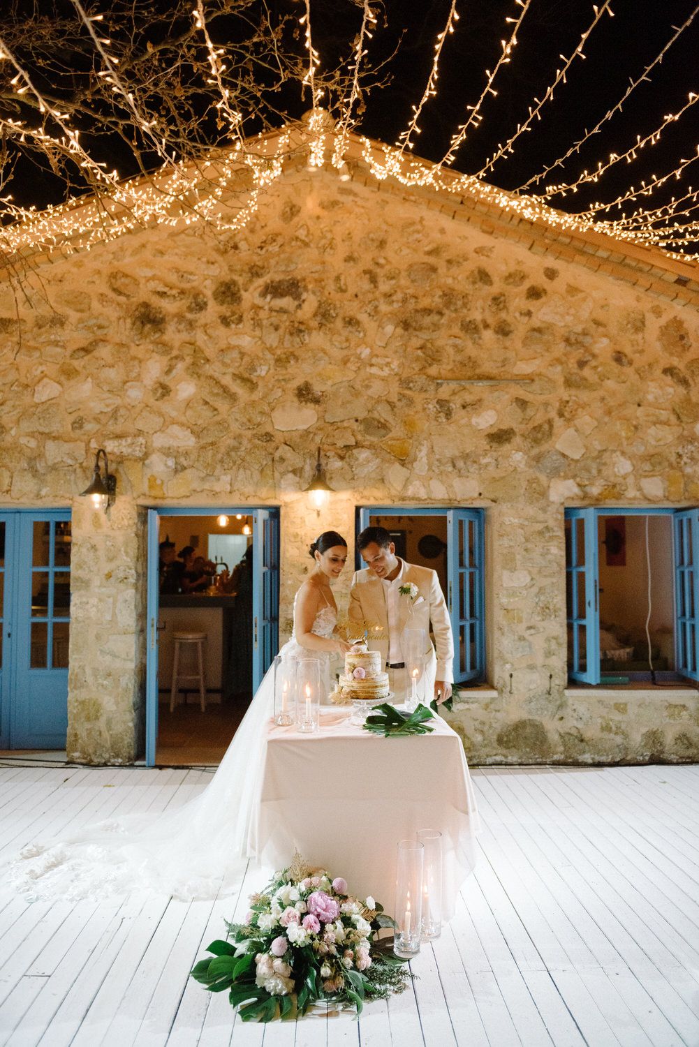 A couple cuts their wedding cake in front of a rustic stone wall adorned with string lights.