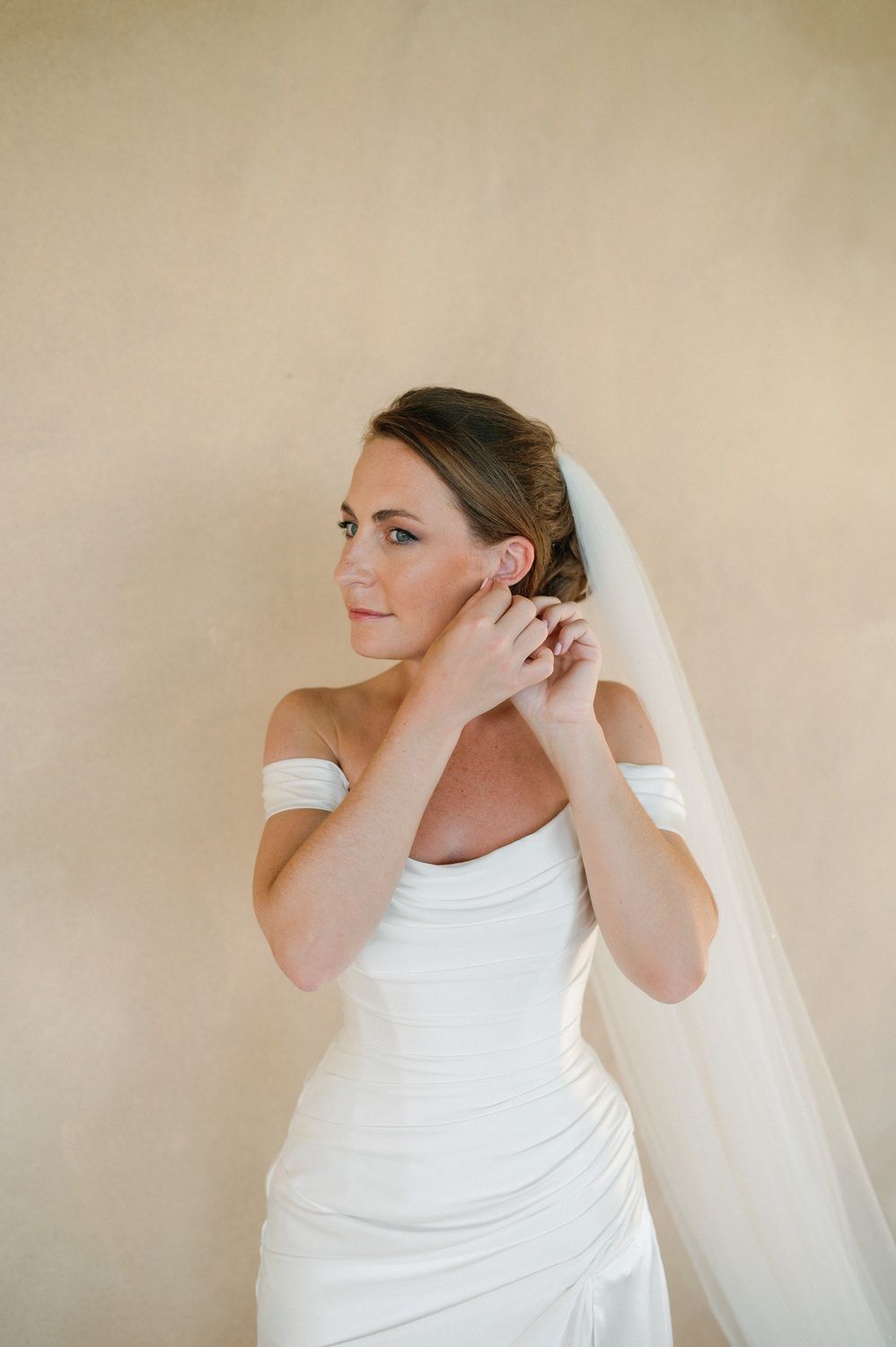 A bride in a white gown adjusts her earring while wearing a flowing veil against a neutral background.