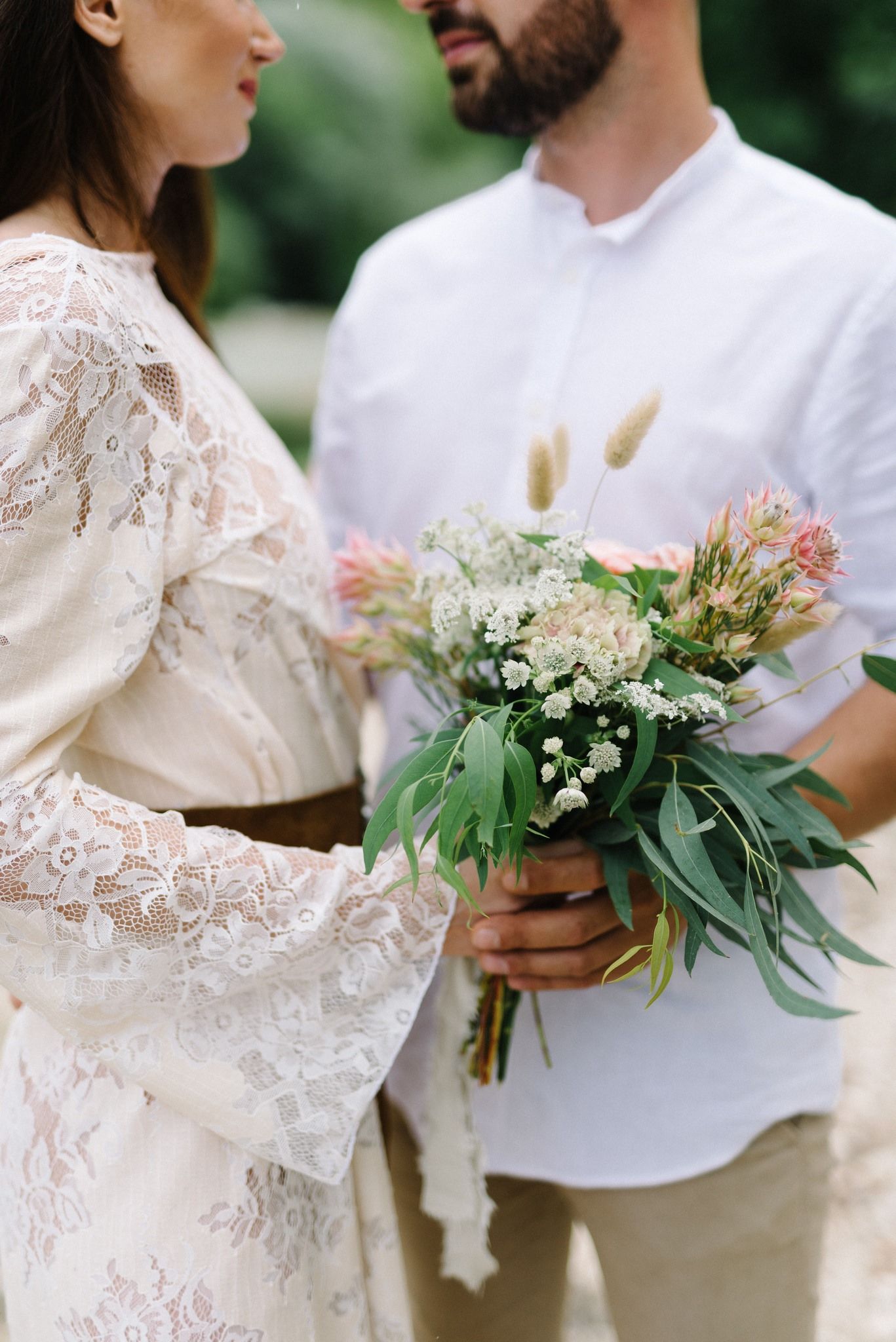 Close-up of couple holding delicate bouquet with white and pink flowers against white and lace attire.