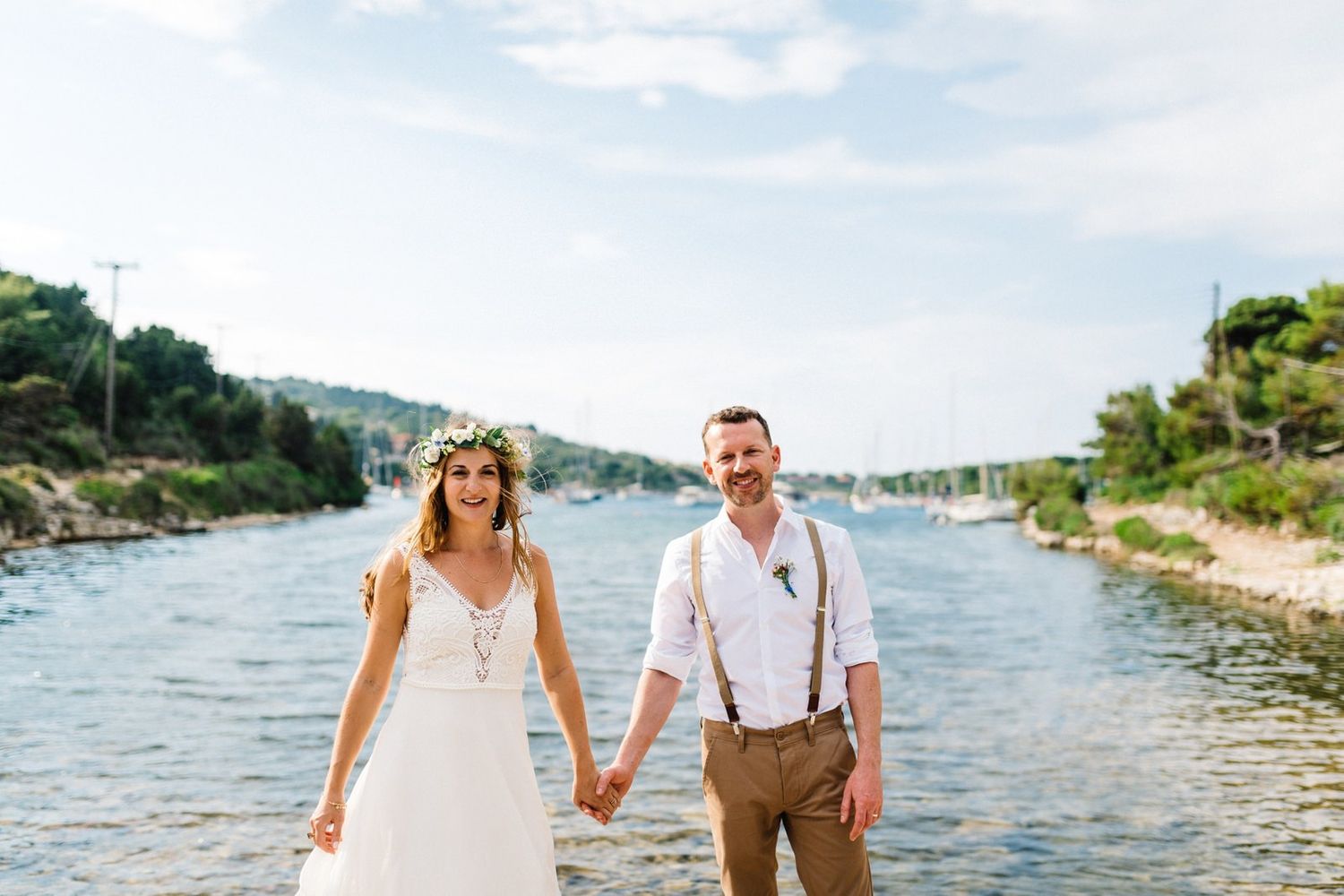 A couple in wedding attire holding hands by a scenic marina with boats and hills in the distance.