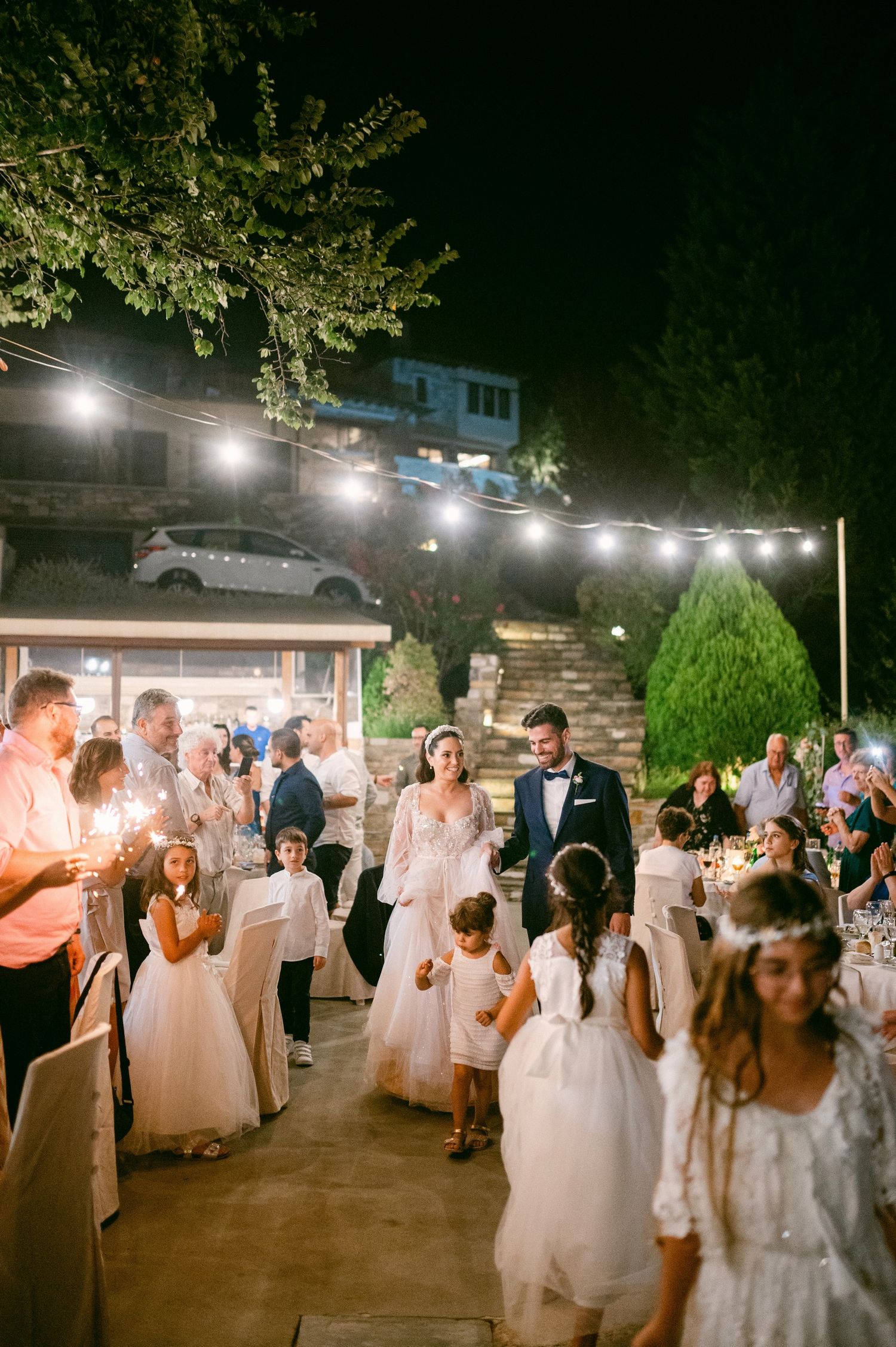 Evening outdoor wedding reception with string lights overhead and guests gathered on a patio under glowing lights.