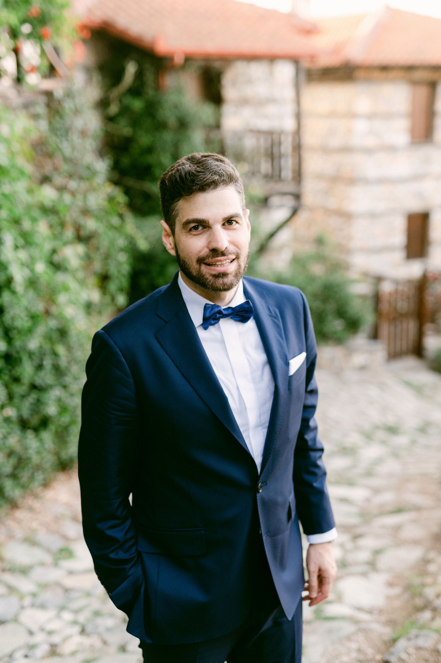 A groom in a navy blue tuxedo with bow tie stands confidently on a rustic stone pathway beside greenery.