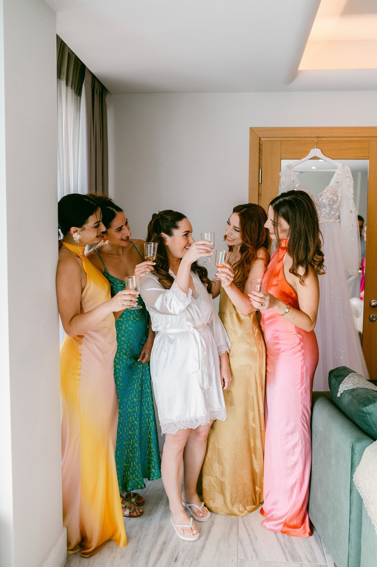 A group in colorful formal dresses gather in a bright hotel room with a curtained window and hangers visible.