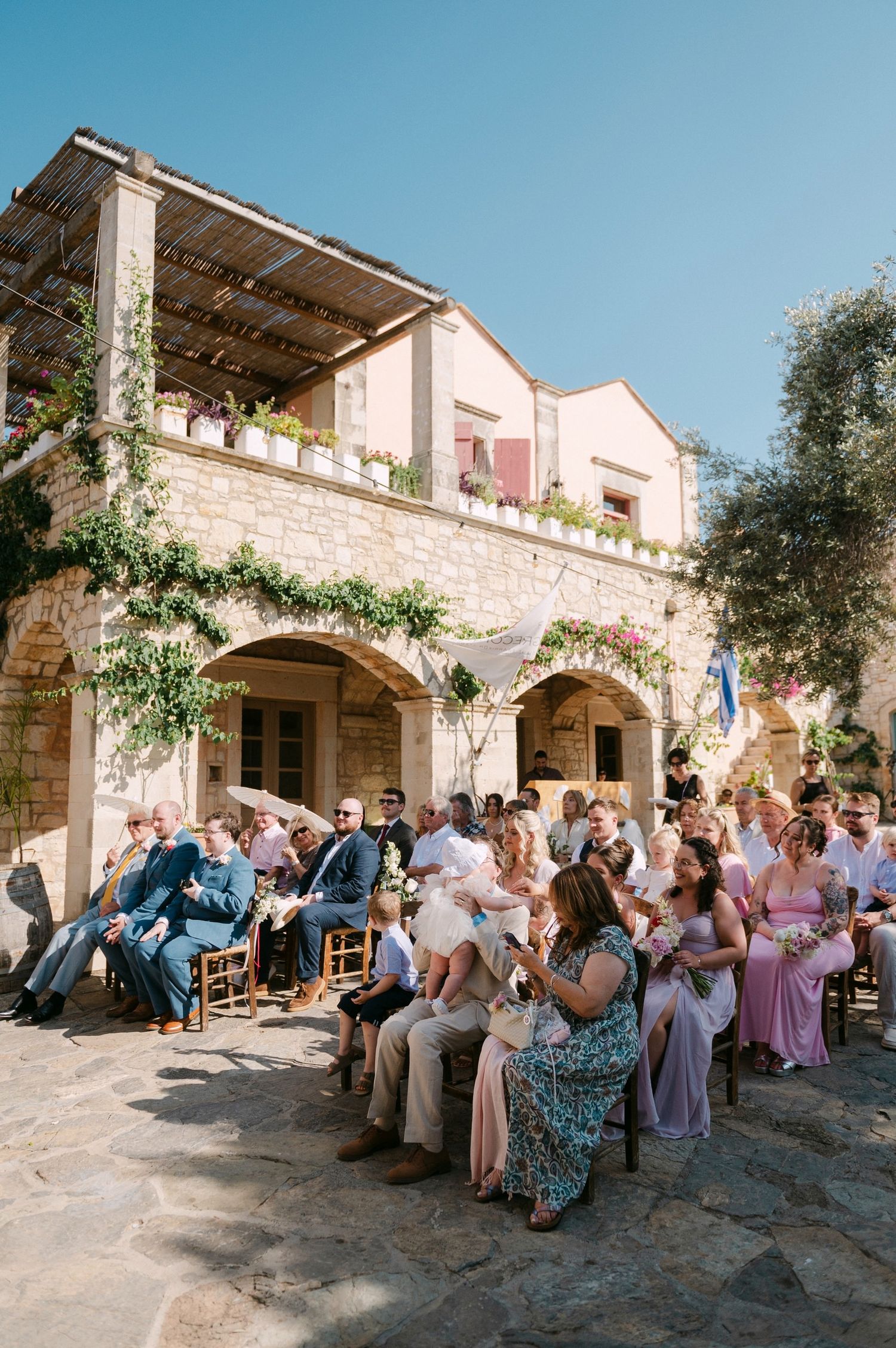 Wedding guests seated outside on rustic stone patio of Mediterranean villa during outdoor ceremony.