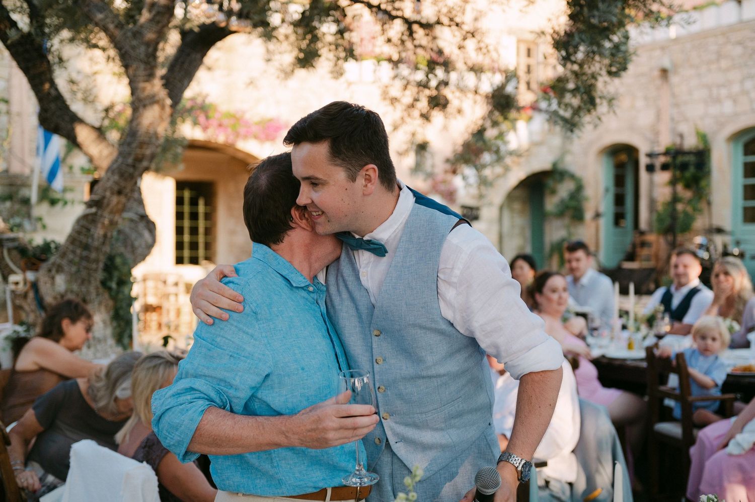 A tender embrace between two people during an outdoor celebration at a rustic venue with guests in the background.