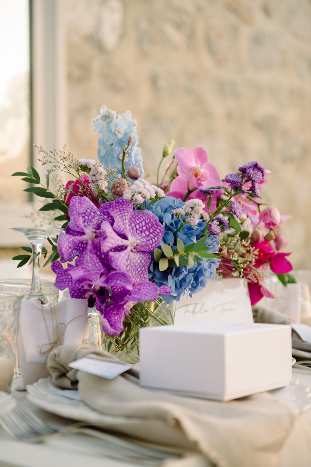 Close-up of purple orchid and blue delphinium floral arrangement with white place setting.
