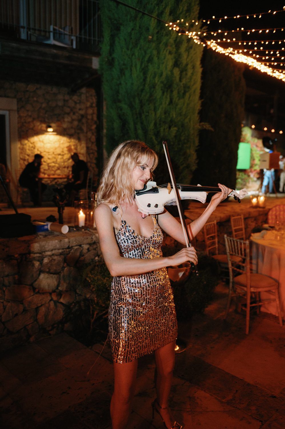 Violinist in a sparkly dress performs at an evening outdoor event under string lights.