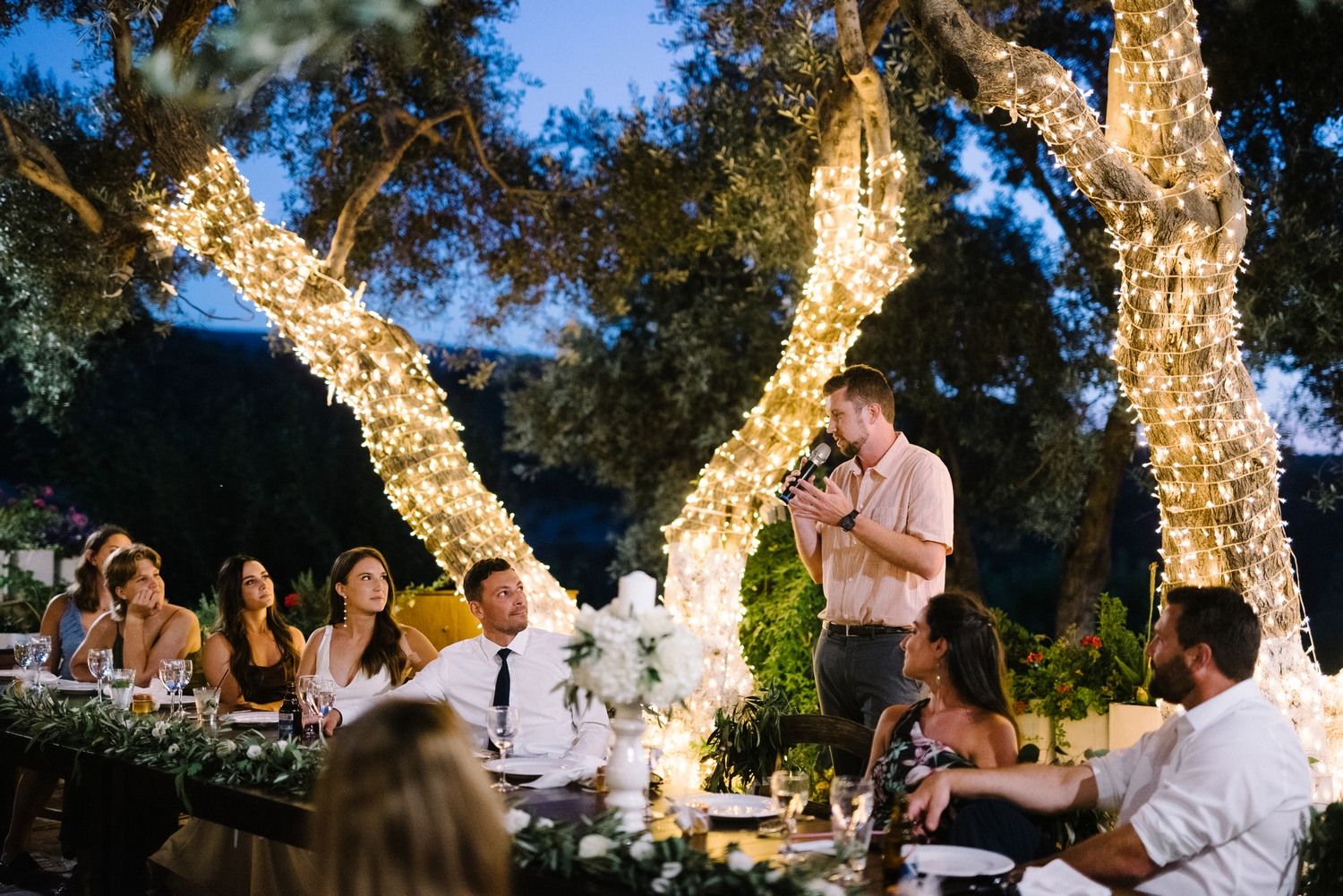 Evening wedding reception setup outdoors with trees wrapped in glowing fairy lights.