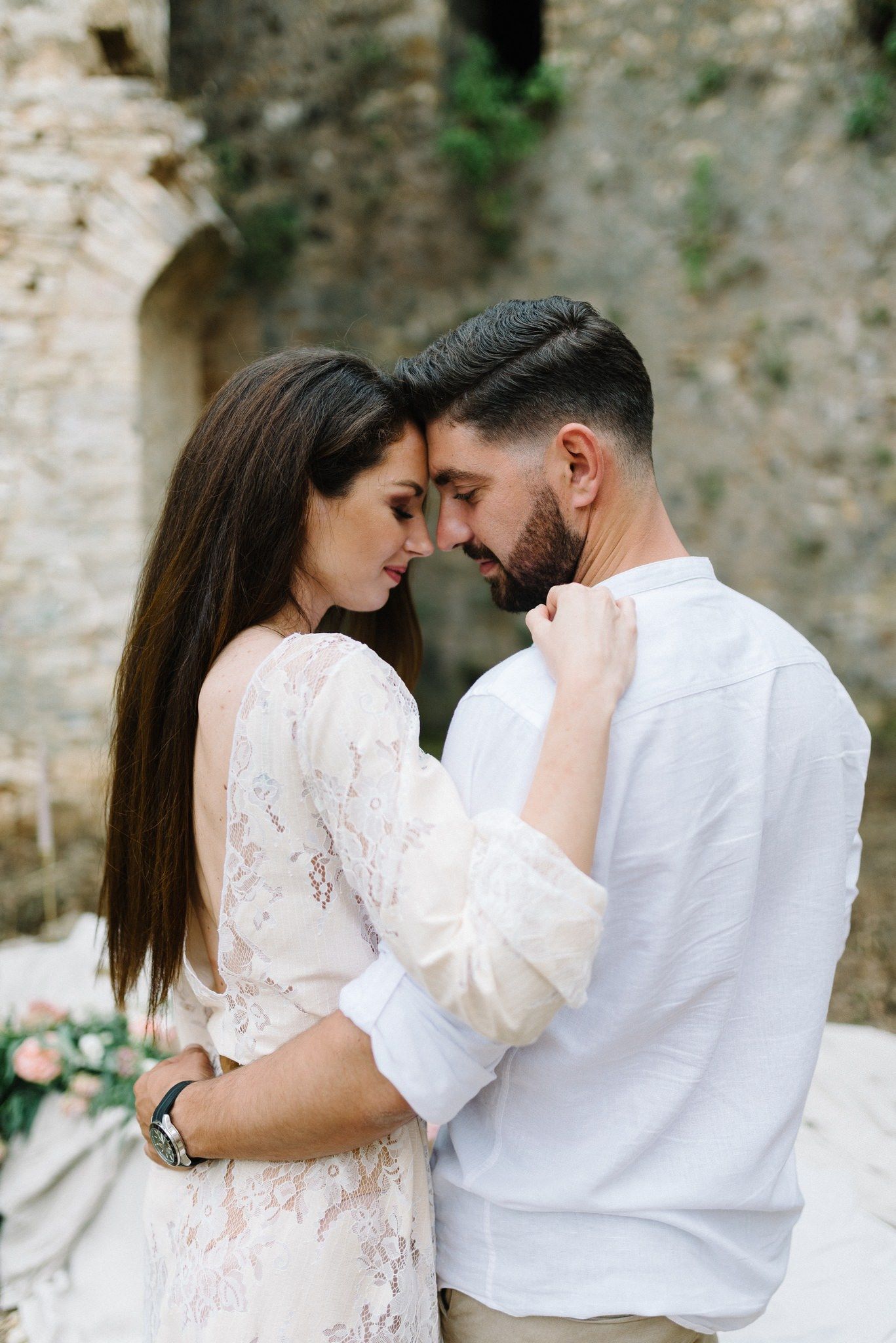 Romantic portrait of couple in white attire against stone wall backdrop.