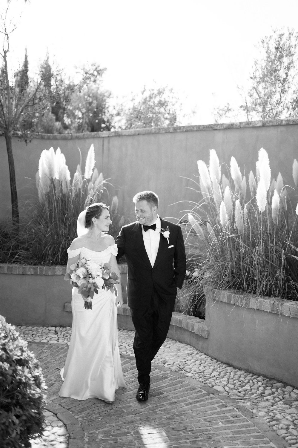 A black and white wedding photograph showing a couple walking along a brick path with ornamental grasses in the background.