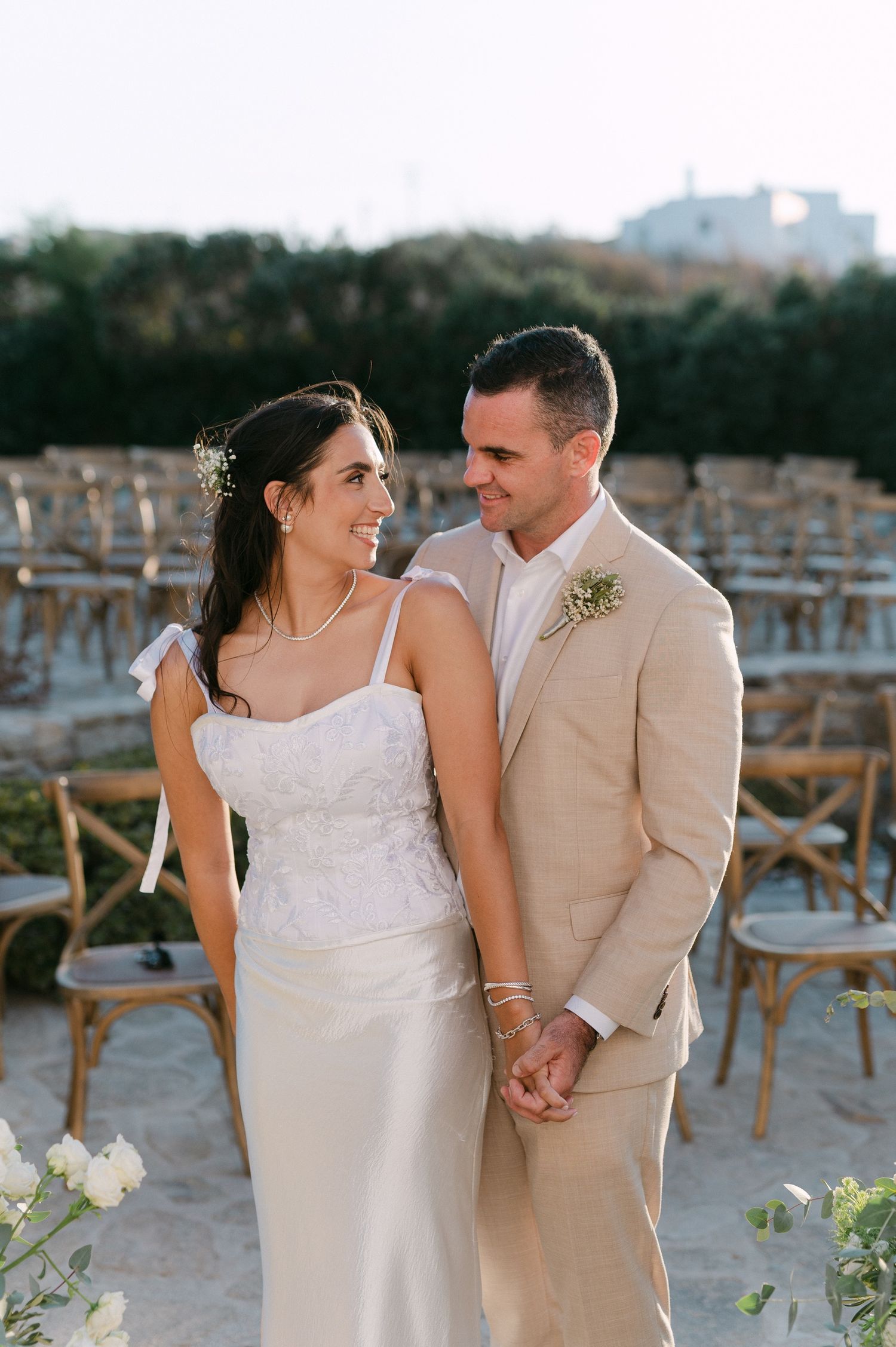 A happy couple shares intimate moments on a beach during their wedding photoshoot with rustic wooden chairs in the background.