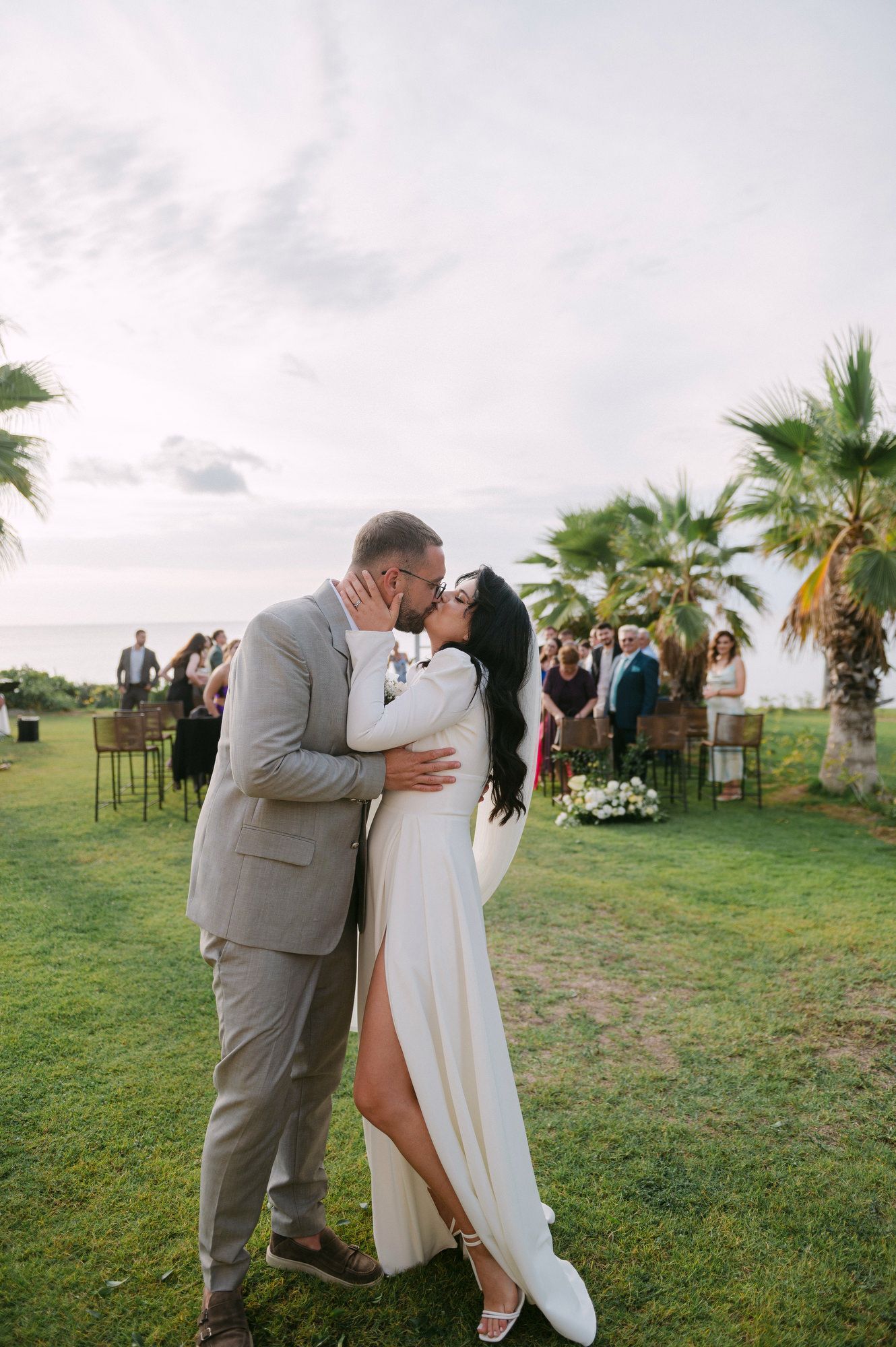 A romantic outdoor wedding ceremony on a grassy lawn with palm trees and guests watching in the background.