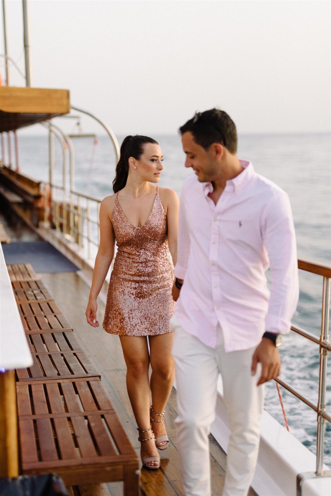 Couple in pink sequin dress and white outfit walking along wooden dock by ocean at sunset.