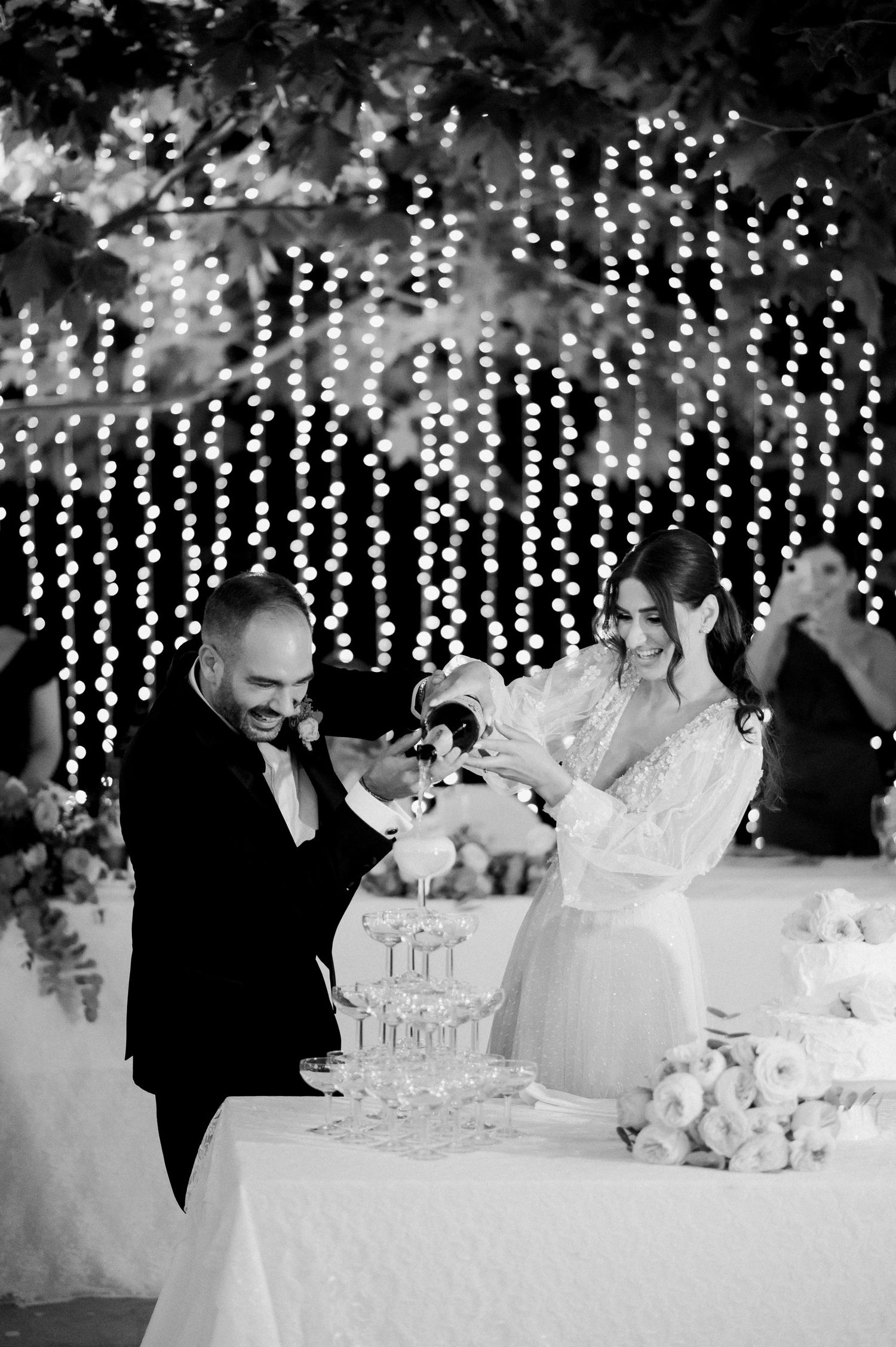 A couple pours champagne into a tower of glasses at their wedding reception with twinkling lights in the background.