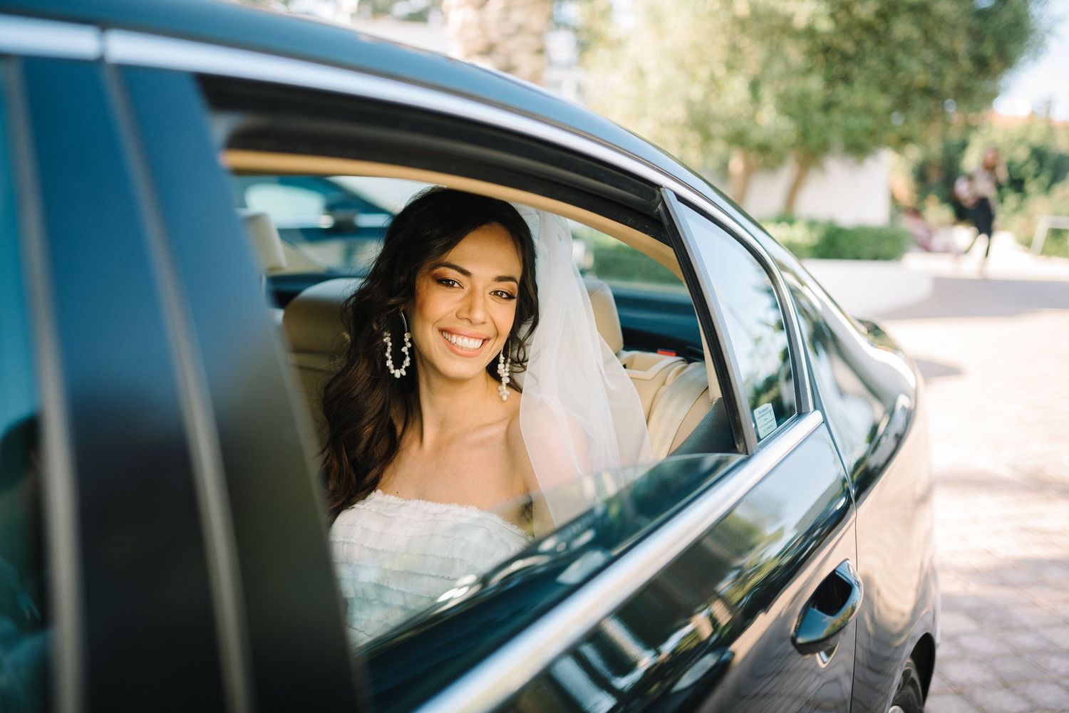 Bride sits in the car while smiling at the camera in a sunny weather waiting to get married