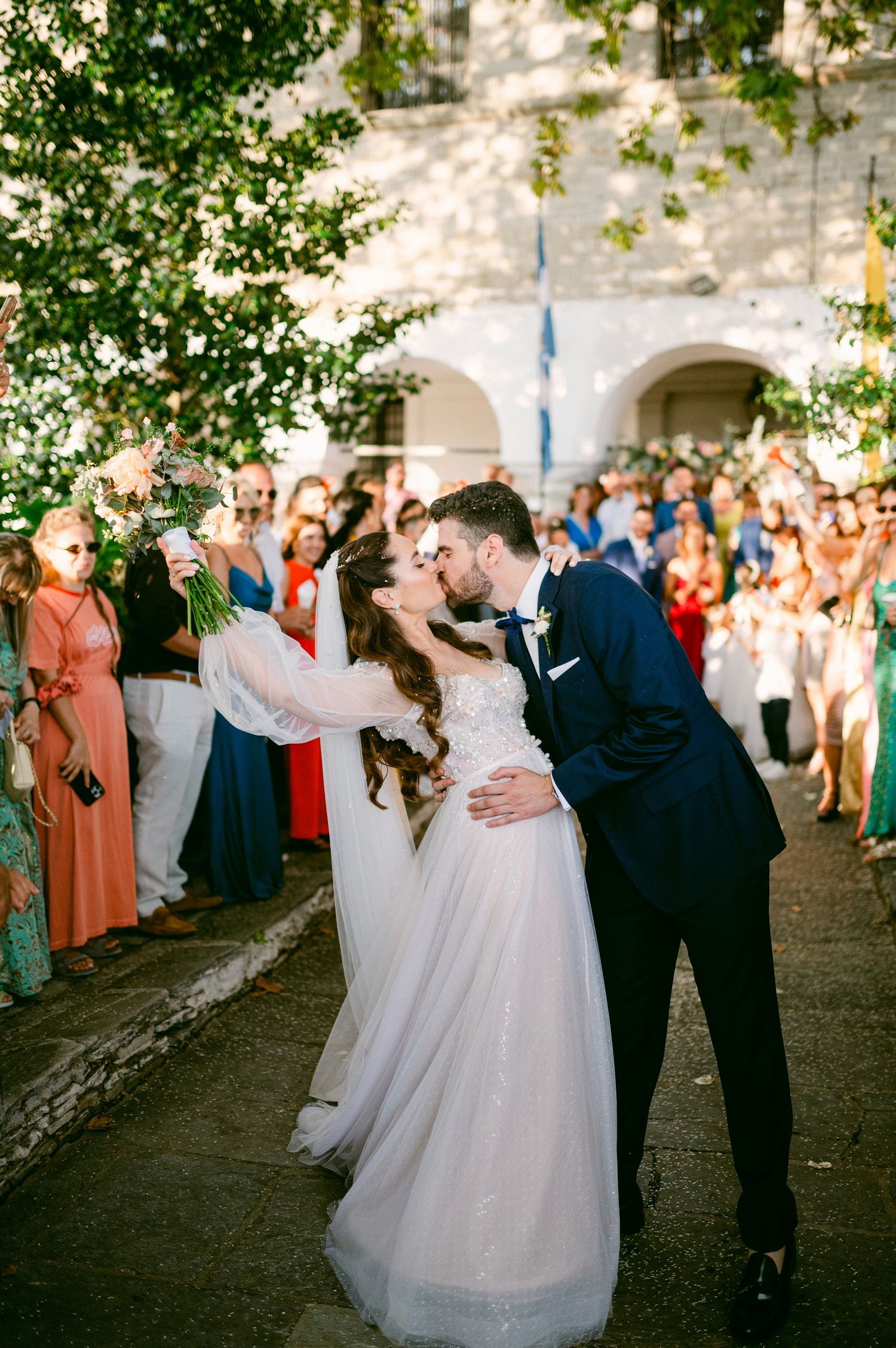A couple shares a romantic dip and kiss at their outdoor wedding celebration while guests cheer in the background.