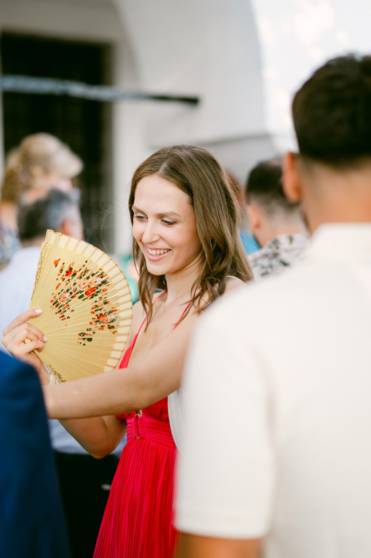A guest in a vibrant red dress smiles while holding a decorative Asian paper fan at a social gathering.