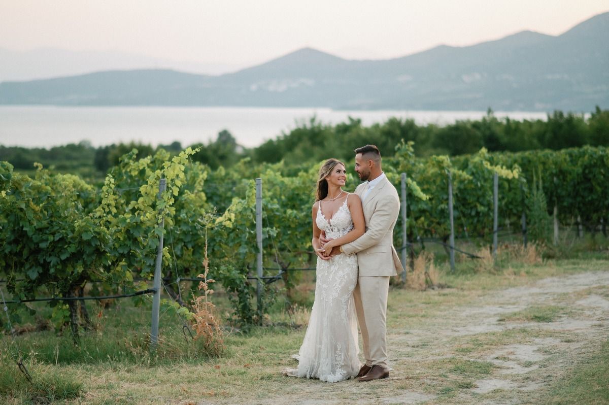 A couple poses in a vineyard with mountains and lake in the background during sunset.
