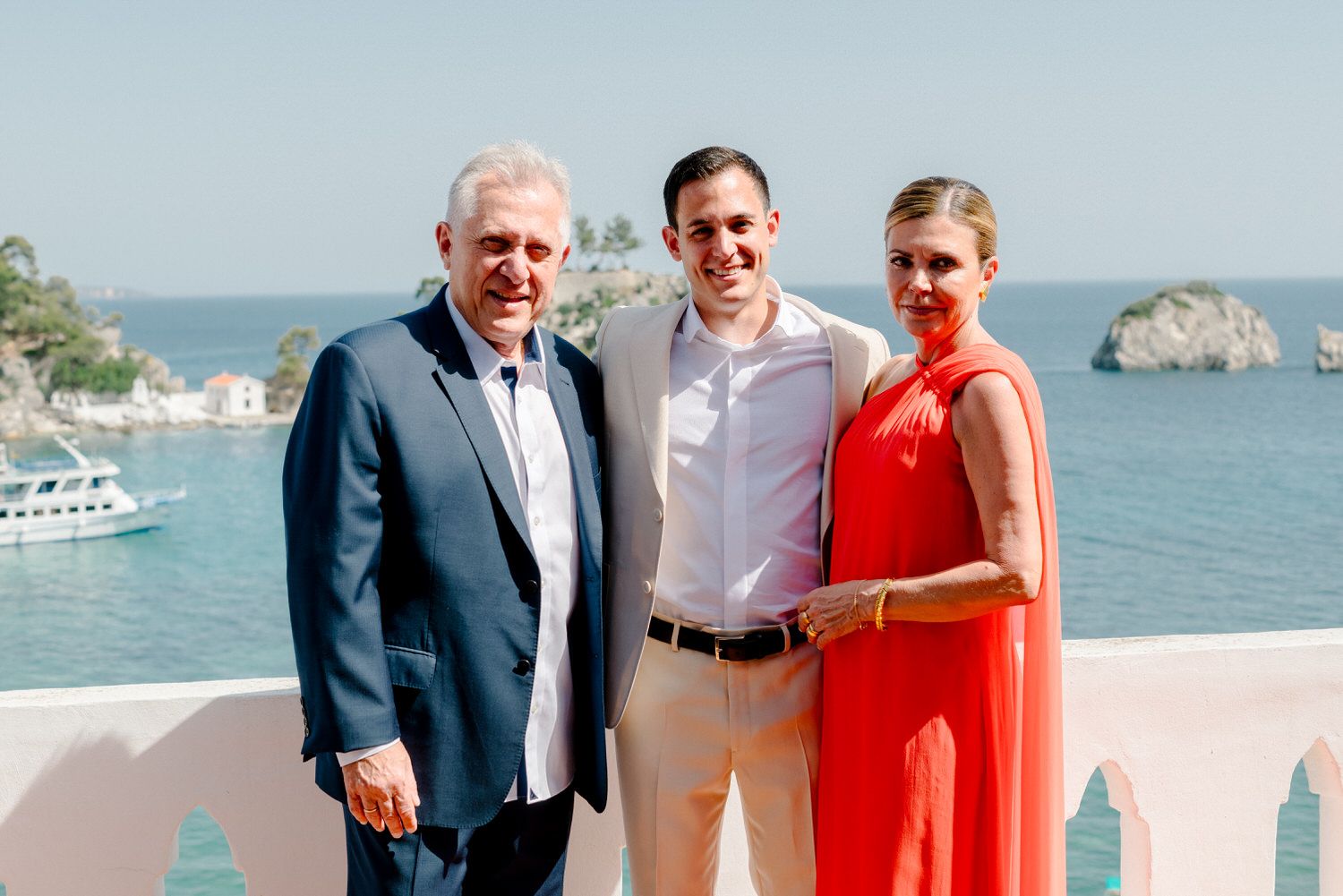 Three people pose on a balcony overlooking a Mediterranean coastline with rocky islands and boats in the background.