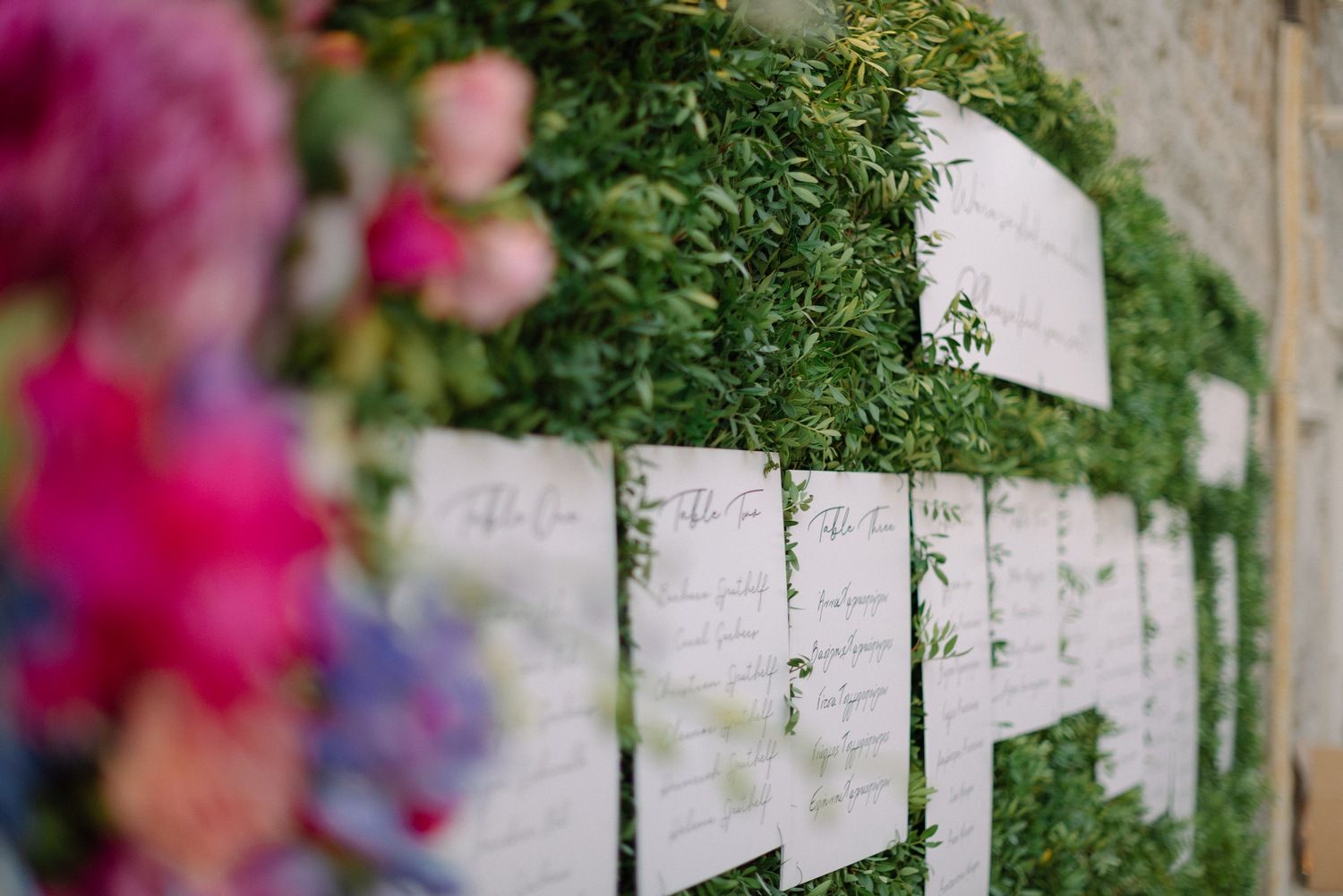 Wedding seating chart display decorated with greenery and pink flowers on a white wall.