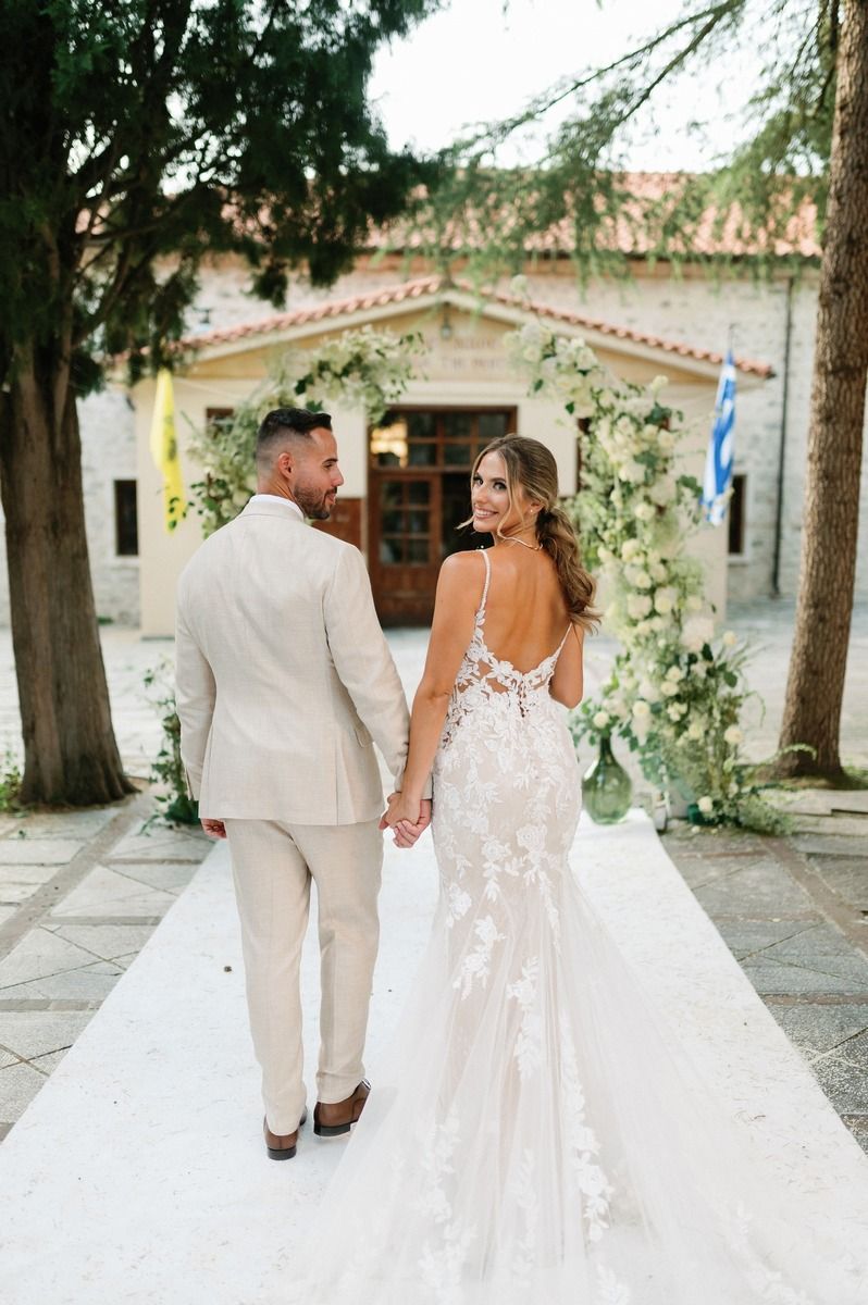 Newlyweds walk hand in hand on white aisle with floral arch and Greek flag visible.
