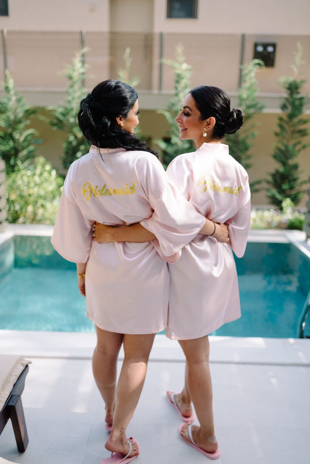 Two people in matching pink robes standing by pool wearing bridesmaid attire.