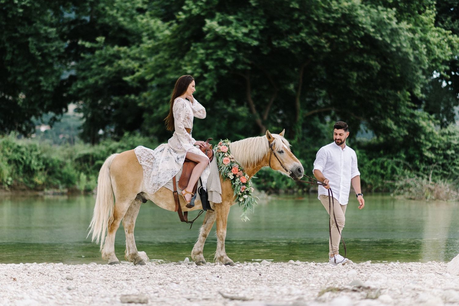 A romantic moment by a lake with a palomino horse on a sandy beach surrounded by lush trees.