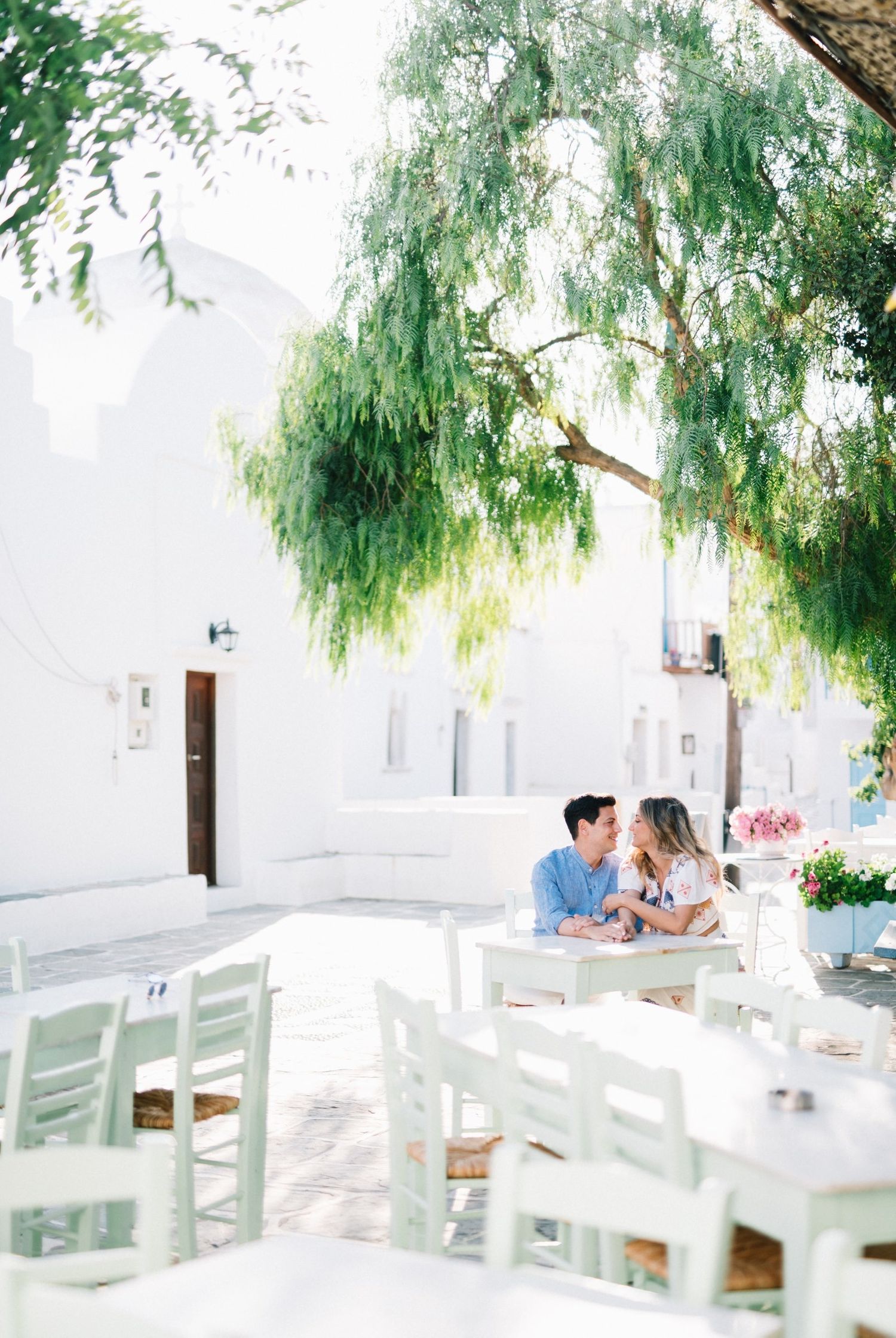 Serene outdoor cafe setting with white chairs and willow trees in Greece.