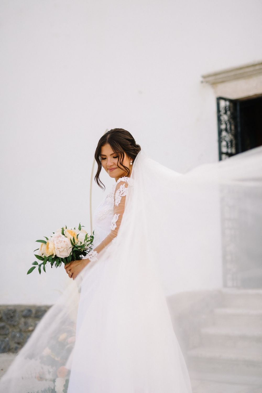 A bride in a flowing white wedding gown and veil poses elegantly against Ypapanti church in Corfu in a romantic bridal portrait.
