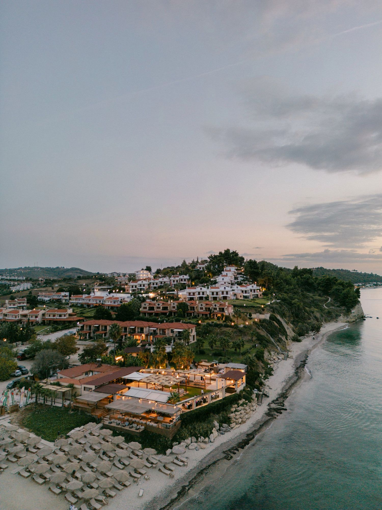 Aerial panoramic view of a coastal resort with beach umbrellas and Mediterranean-style buildings at sunset.