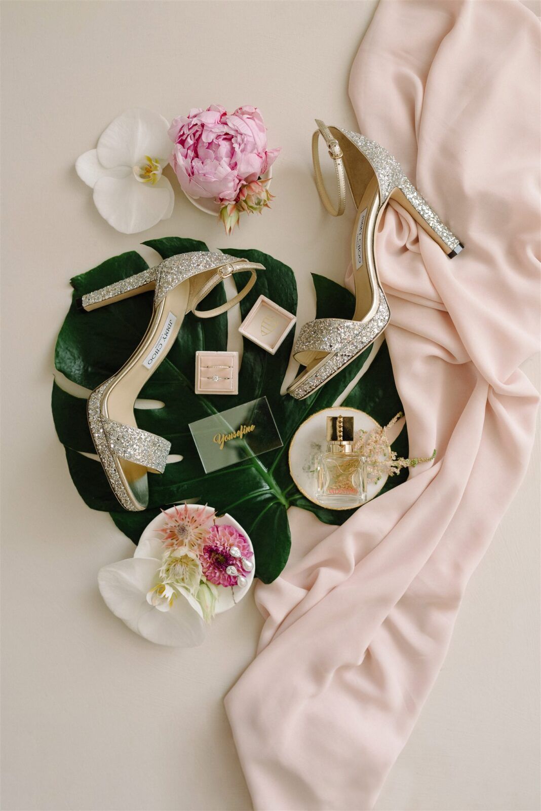 Elegant bridal shoes with sparkly straps arranged with pink flowers, white orchids, and blush fabric on a light background.