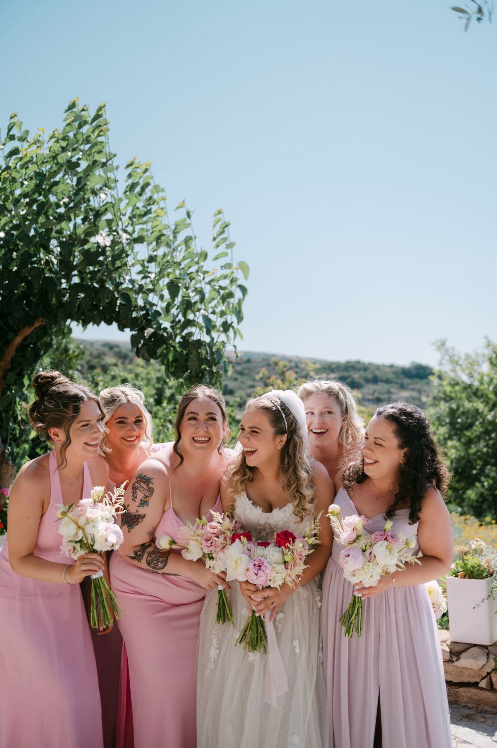 A group of bridesmaids in pink and white dresses laugh togetherat Agreco farms, Crete