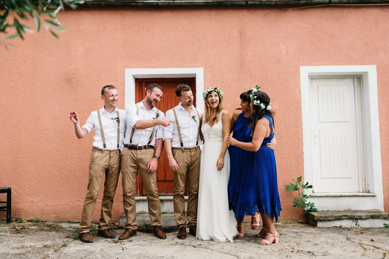 Wedding party posing playfully in front of a coral-colored building with white doors.