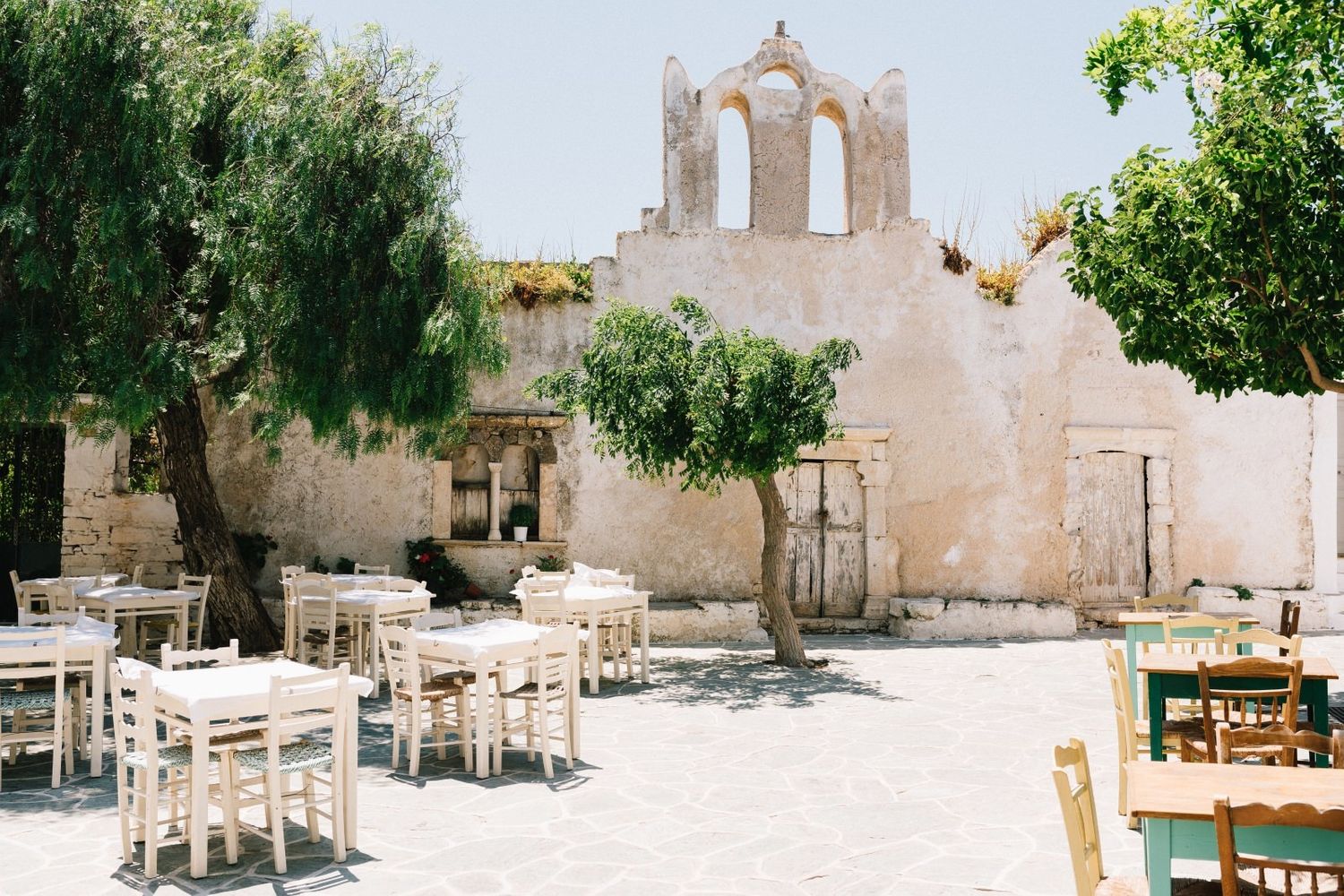 A rustic Mediterranean restaurant courtyard with white tables and chairs in front of a traditional whitewashed church facade.
