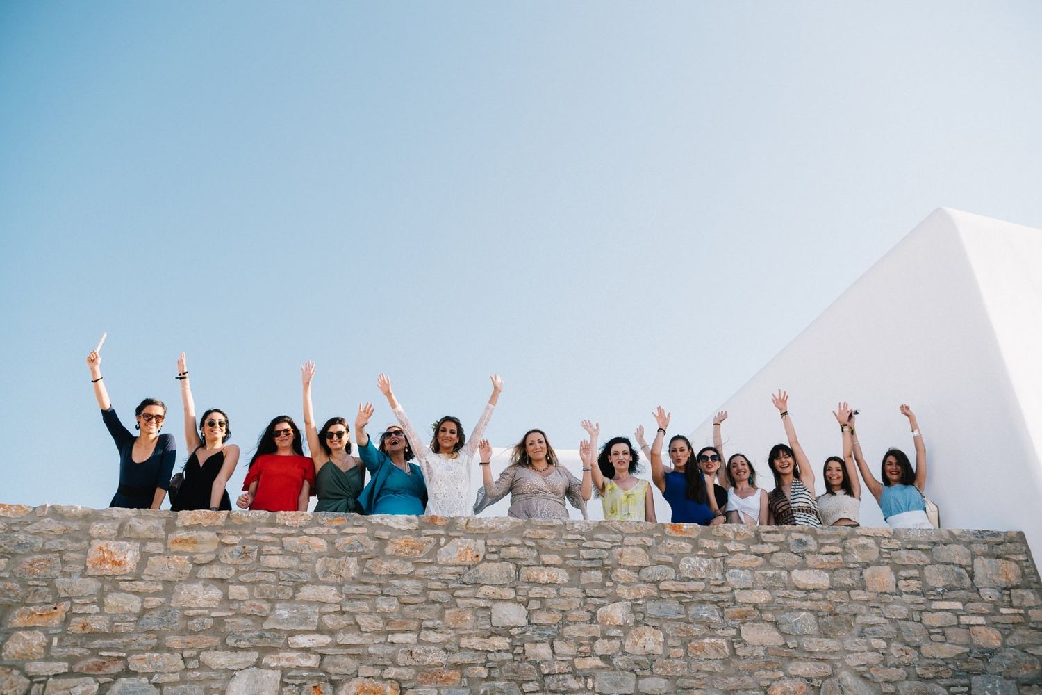 Bridal party celebrating with raised arms on stone wall against bright blue sky.