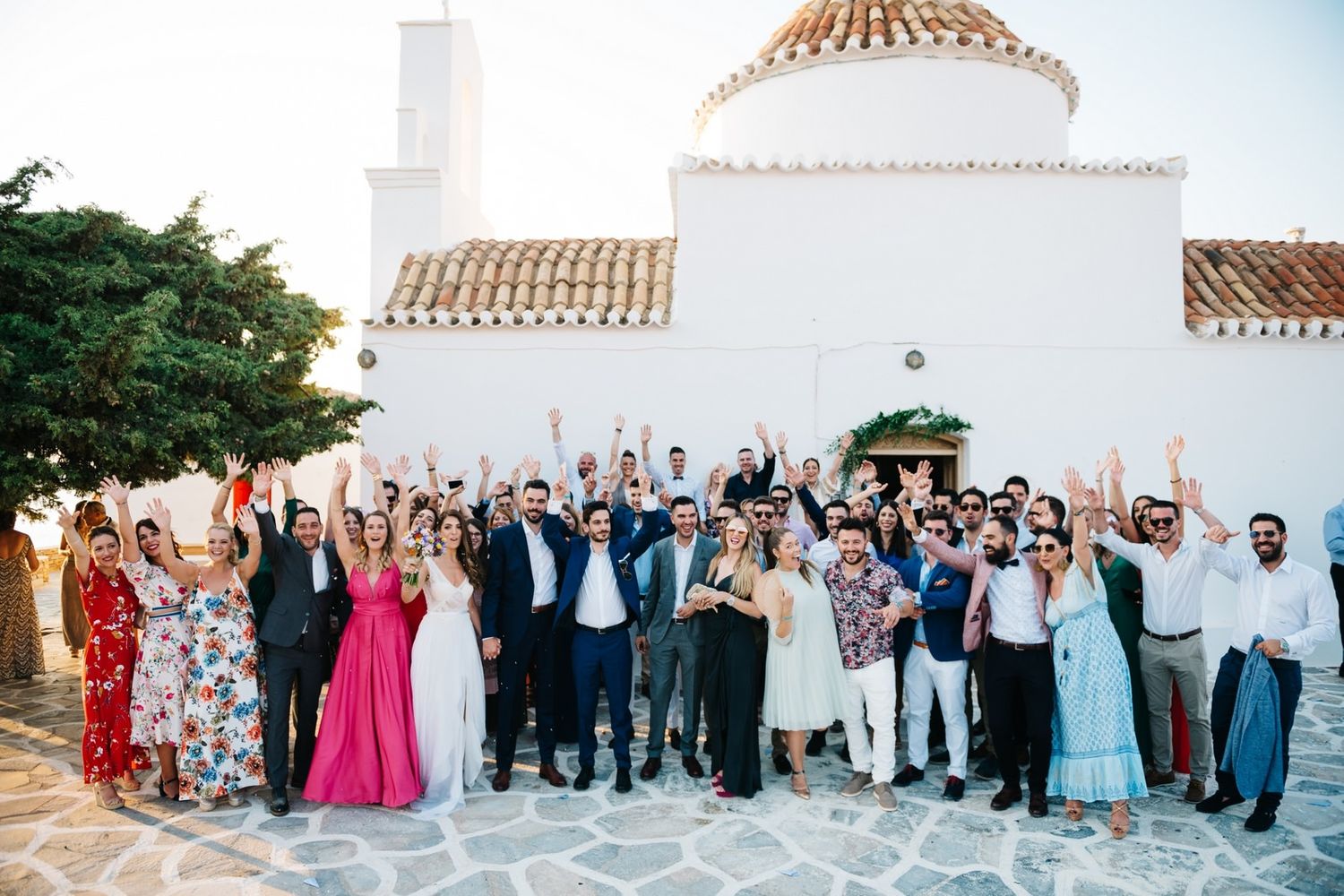 Group celebration photo outside white Mediterranean church with jubilant wedding guests.