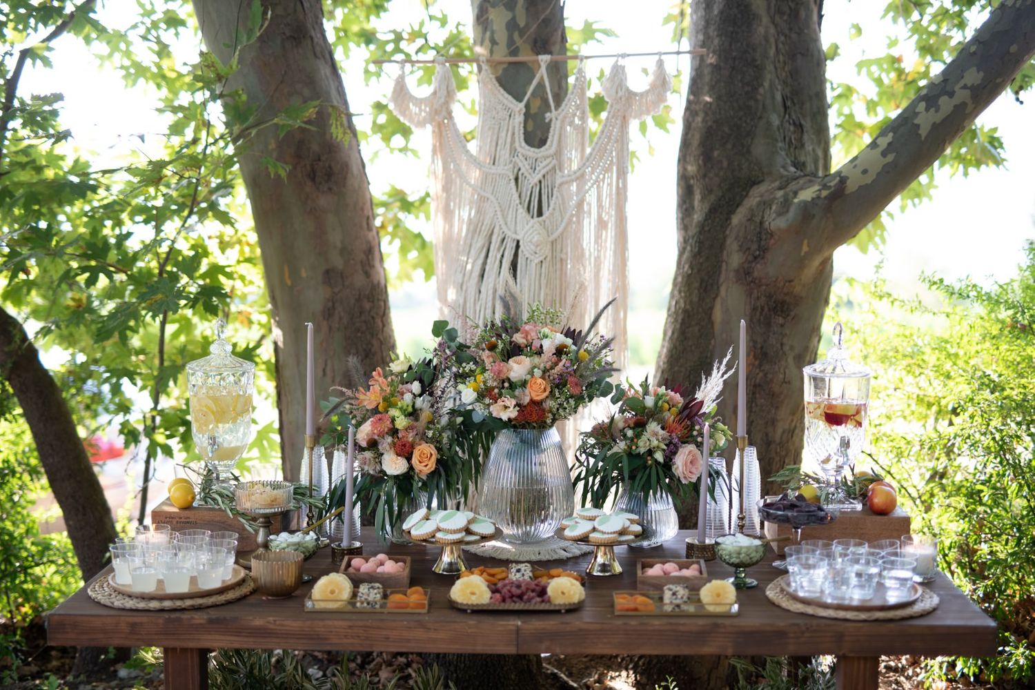 Rustic outdoor wedding table setup under trees with floral centerpieces and hanging decorations.