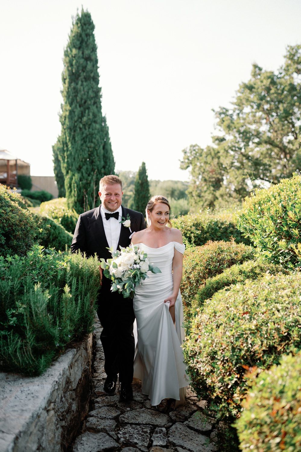 Wedding couple walking along stone pathway surrounded by manicured garden and cypress trees.