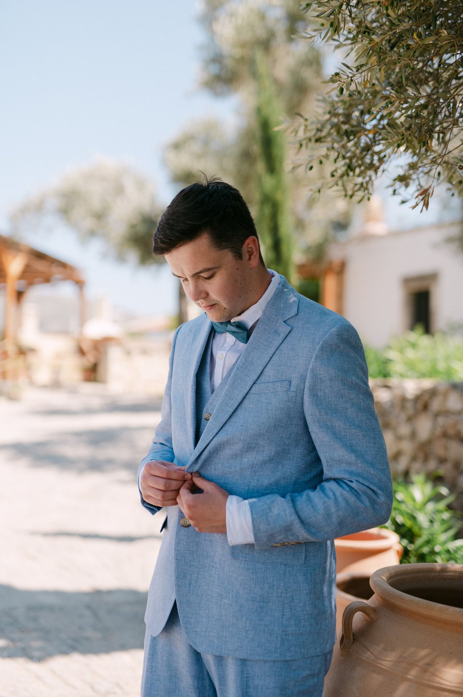Someone in a light blue suit adjusts their jacket button while standing outside on a sunny day.