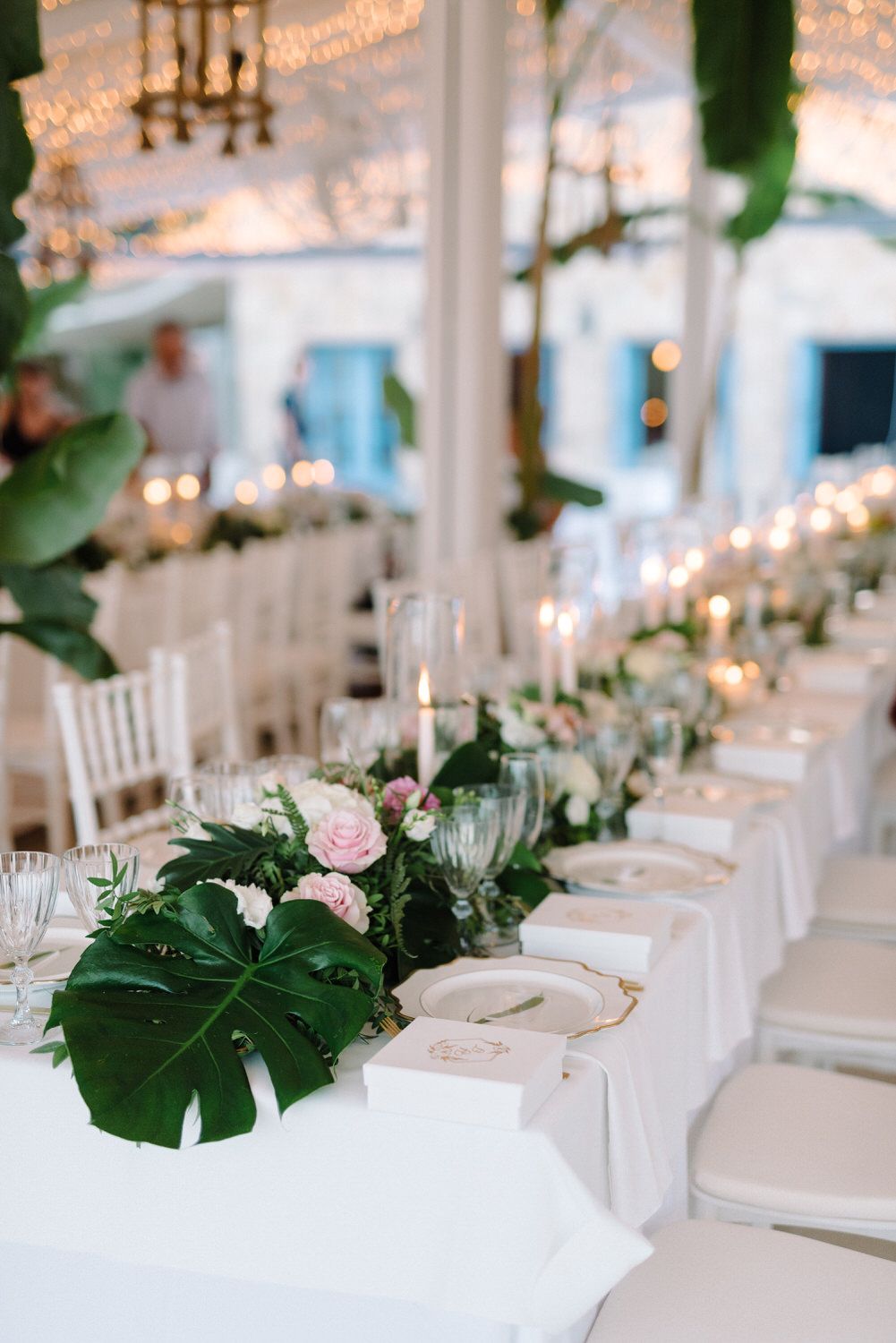 White wedding reception tables adorned with monstera leaves, roses, and glass candleholders.