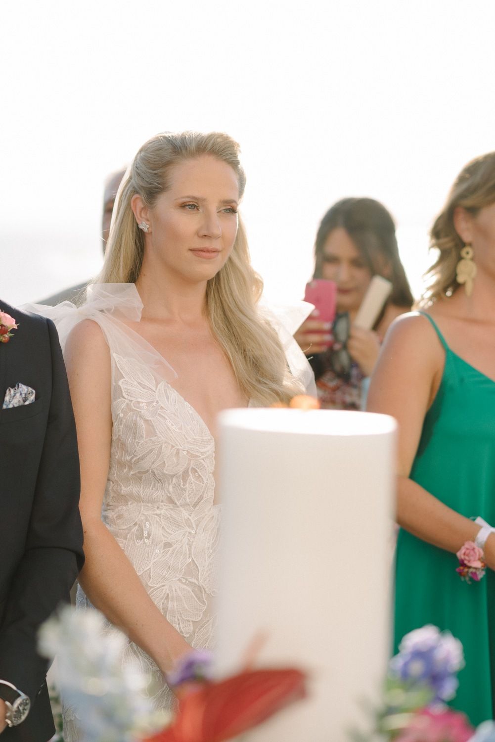Elegant bride in lace gown at outdoor ceremony with guests in background.