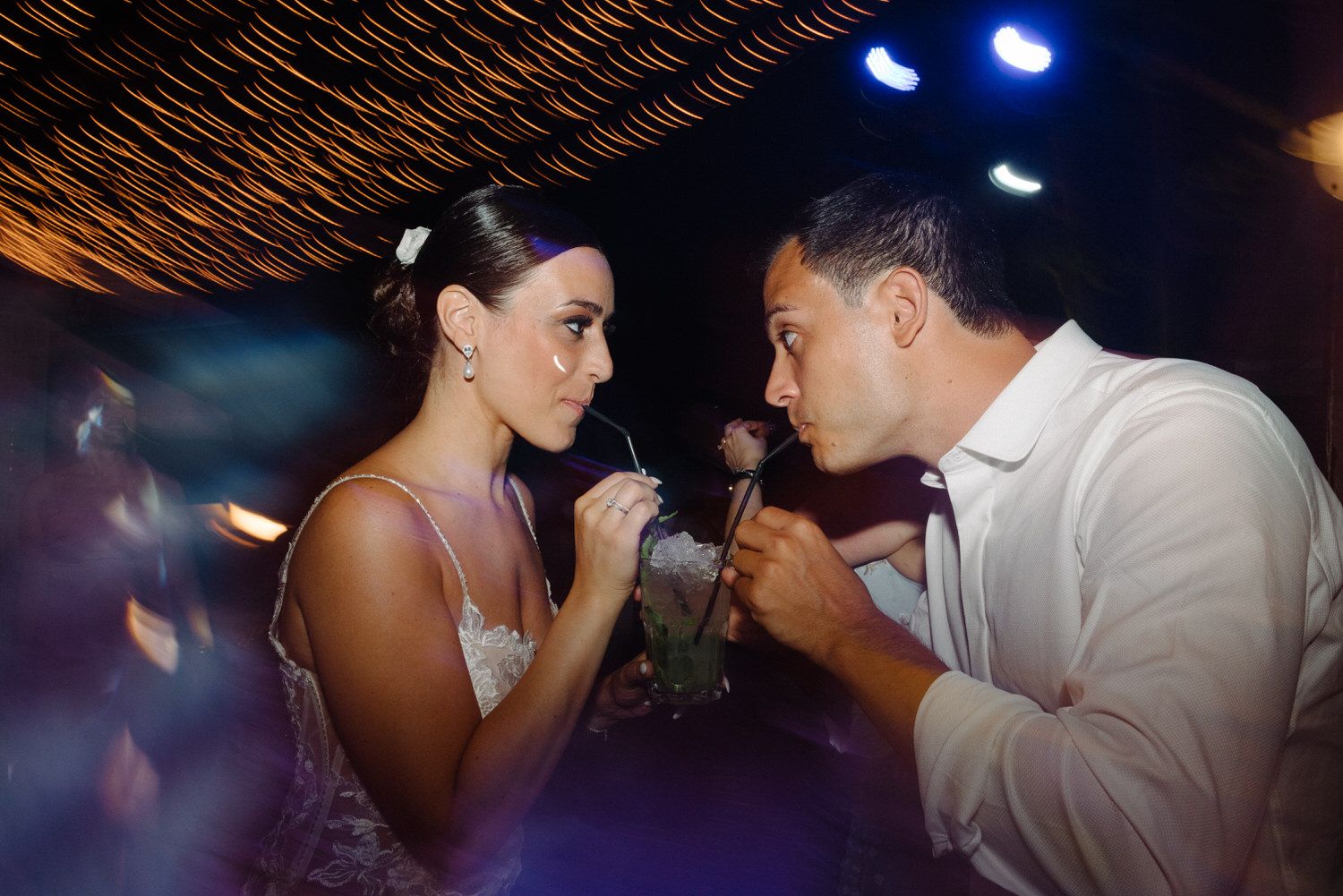 A couple shares drinks during their wedding reception under atmospheric lighting.