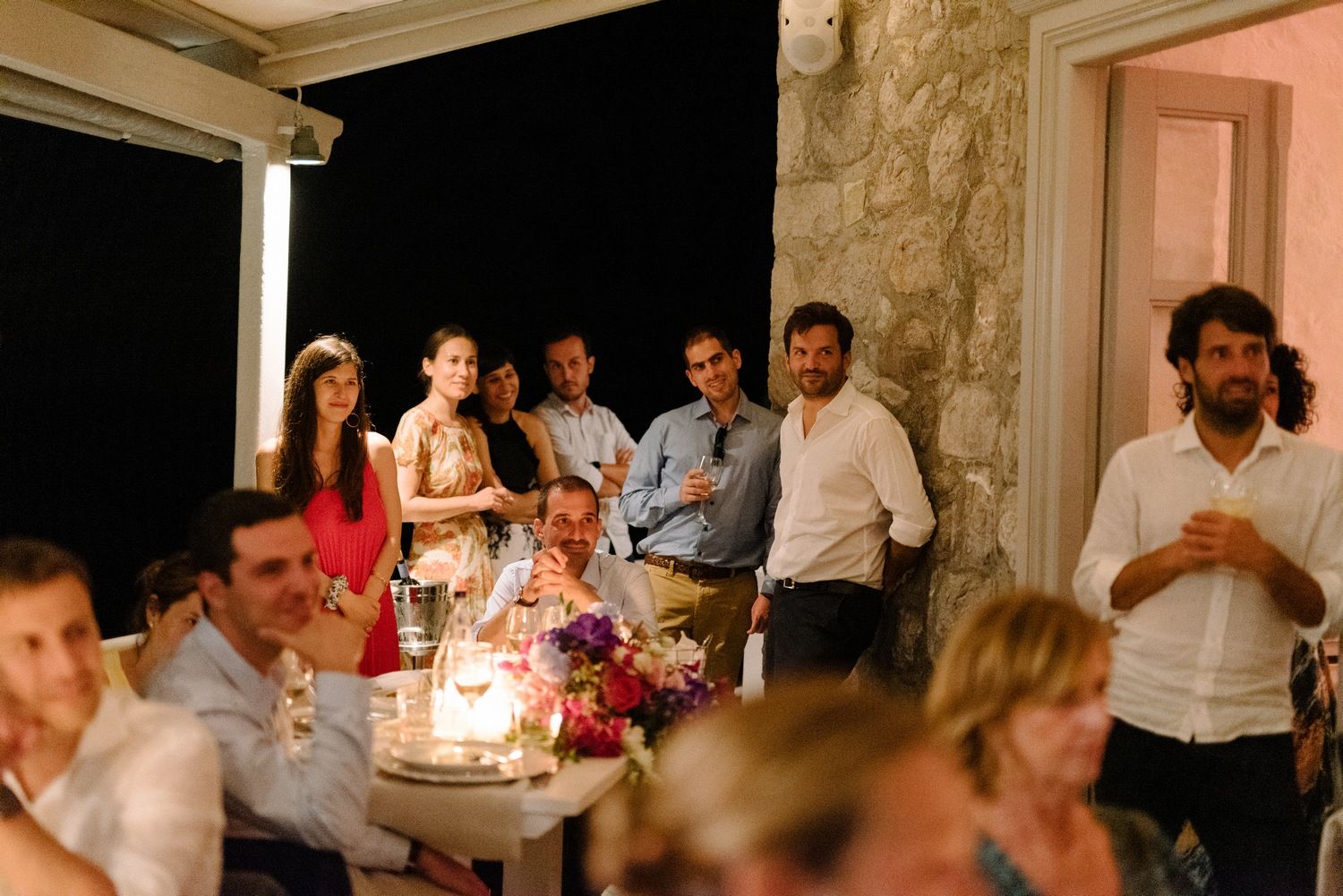 Wedding guests gathered around candlelit tables at an evening celebration with stone wall backdrop.