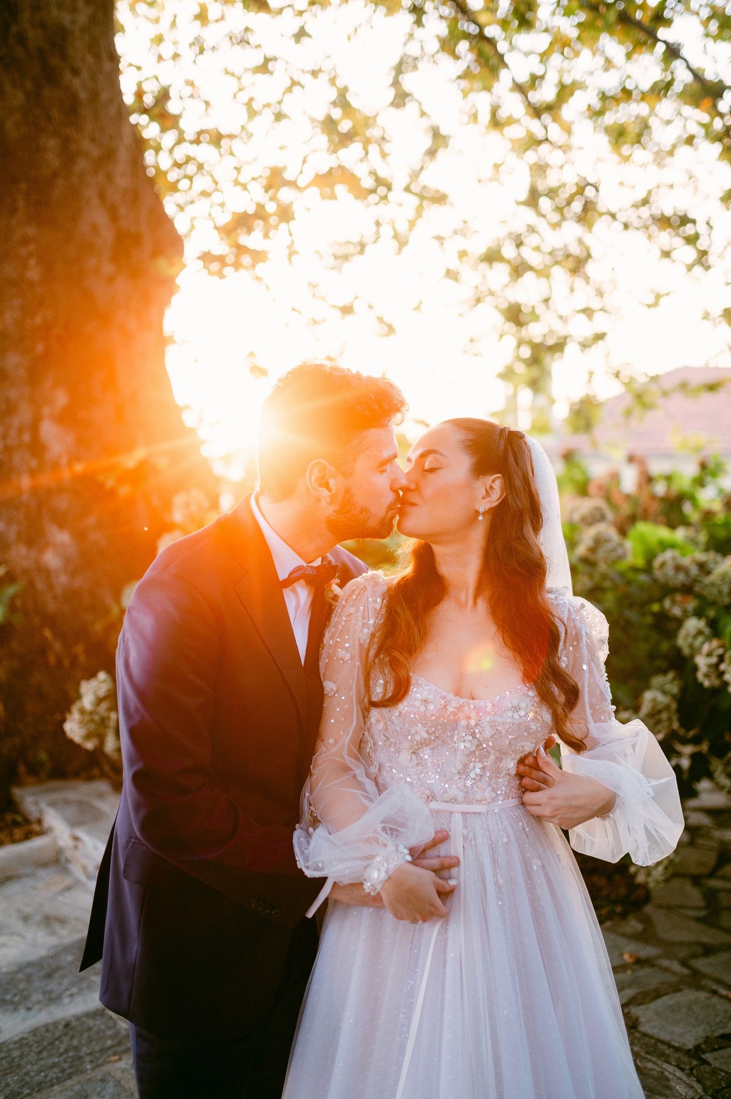 A romantic bride and groom share a kiss at sunset with golden sunlight streaming through the trees.