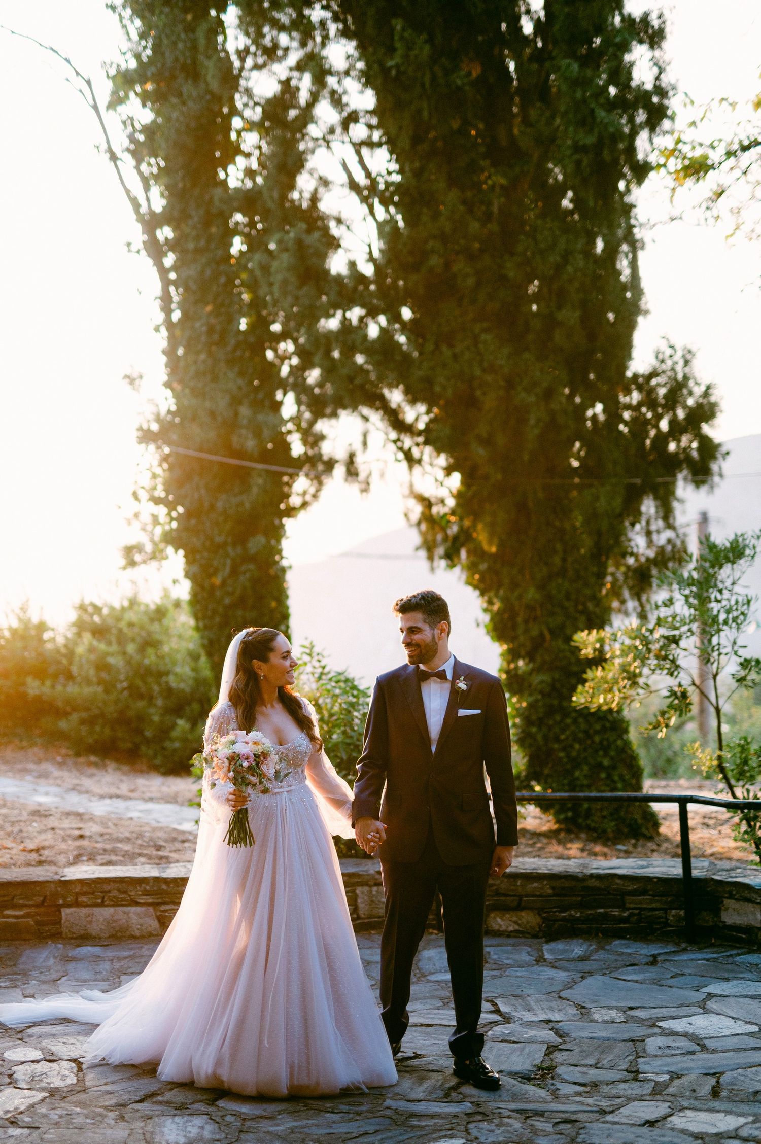 Newlyweds walking together on stone pathway at sunset with tall cypress trees in romantic outdoor setting.