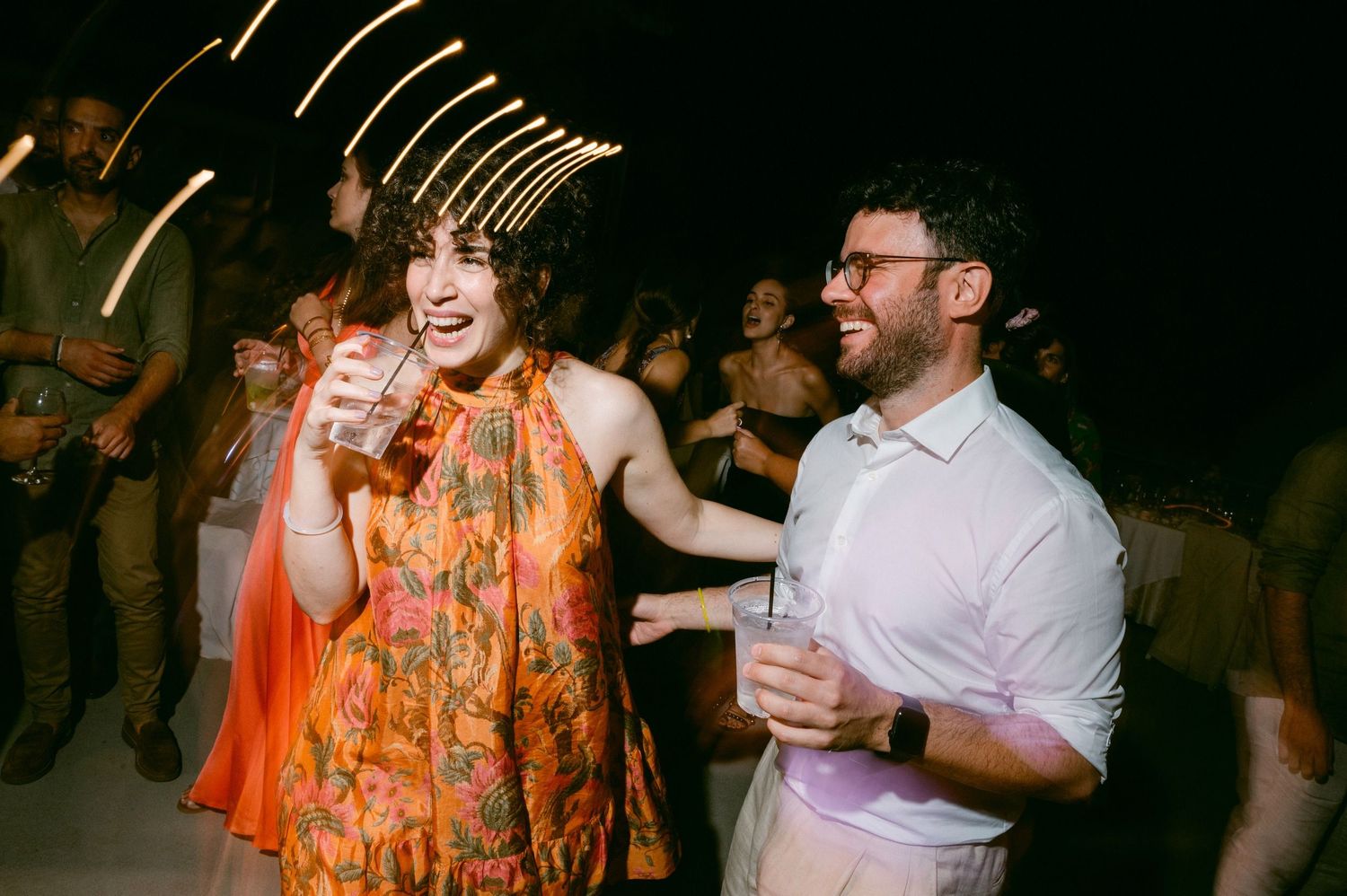 Two people dance together at a nighttime party, one wearing a bright orange floral dress while holding drinks.