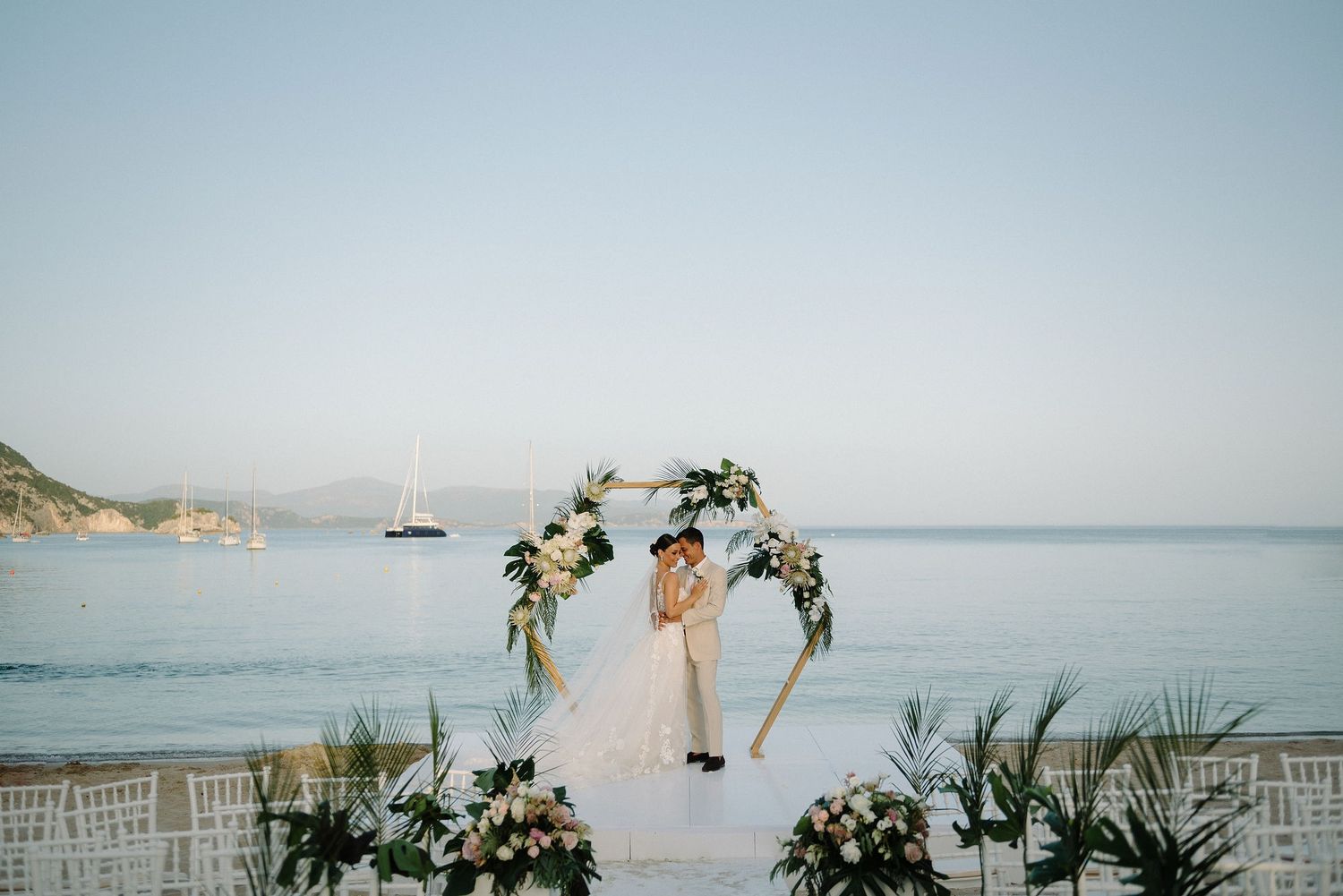 A romantic beachside wedding ceremony setup with floral arch and chairs overlooking a calm ocean bay with sailboat.